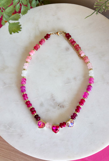 Warm shades of pink and shimmering details make this handcrafted necklace a standout accessory for every season! Featuring beautiful pink beads in a variety of tones accented with gold details, this elegant piece is finished with sparkling heart-cut crystals that add the perfect touch of brilliance. Whether you're dressing up a cozy sweater or adding a feminine touch to your favorite dress, this timeless necklace brings cheerful color and effortless charm to every outfit.