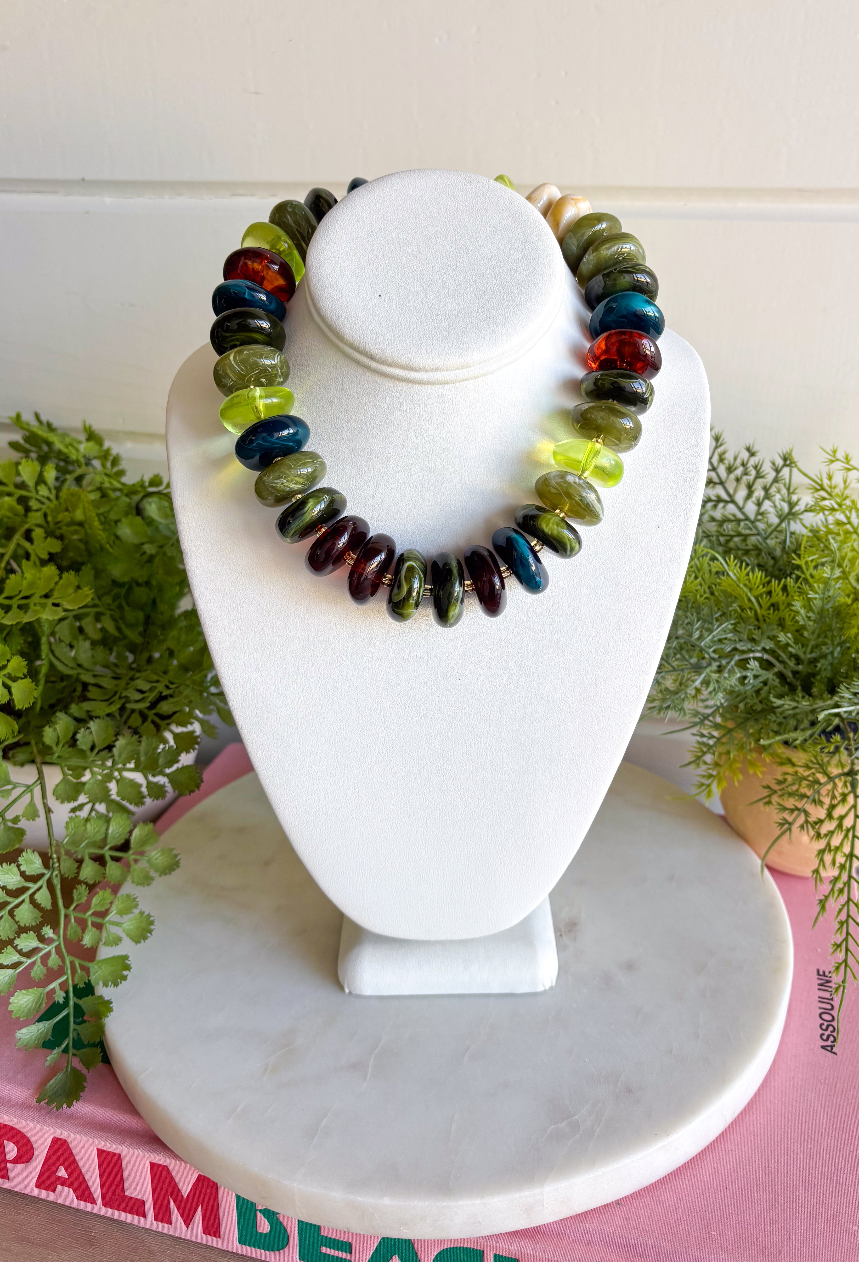 A beaded gemstone necklace displayed on a white jewelry bust. The necklace features a mix of polished round beads in olive green, dark teal, forest green, amber orange, and deep red.