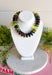 A beaded gemstone necklace displayed on a white jewelry bust. The necklace features a mix of polished round beads in olive green, dark teal, forest green, amber orange, and deep red.