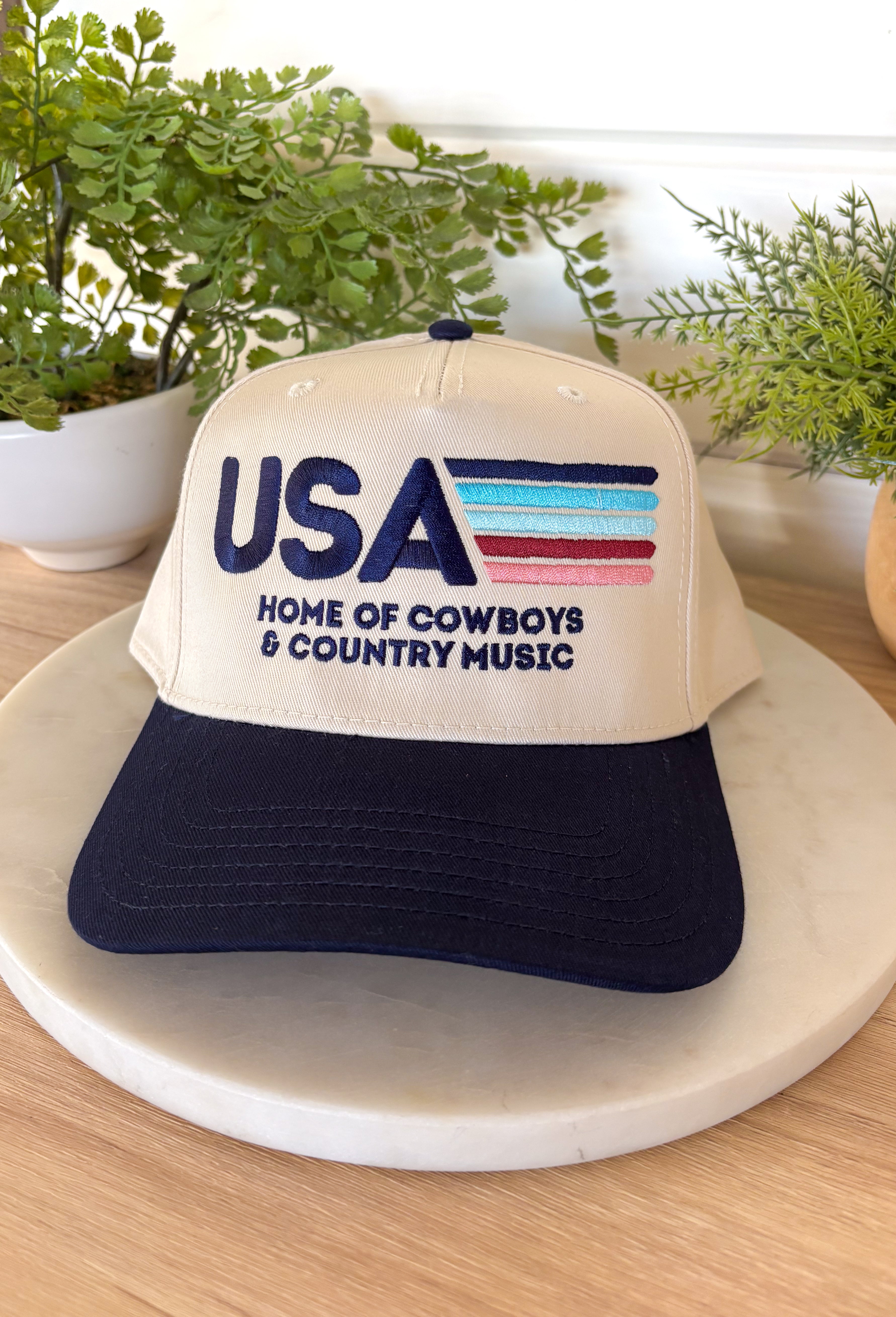 This neutral trucker hat is the perfect laid-back statement for everyday wear. Featuring “USA Home of Cowboys &amp; Country Music” embroidered in navy with colorful stitched accents, it adds a fun pop of detail while keeping things classic and easy to style. The neutral base makes it an effortless match with just about anything, and the adjustable back ensures a comfortable, just-right fit. Whether you’re running errands, heading to a concert, or finishing off a casual outfit, this hat brings a little perso