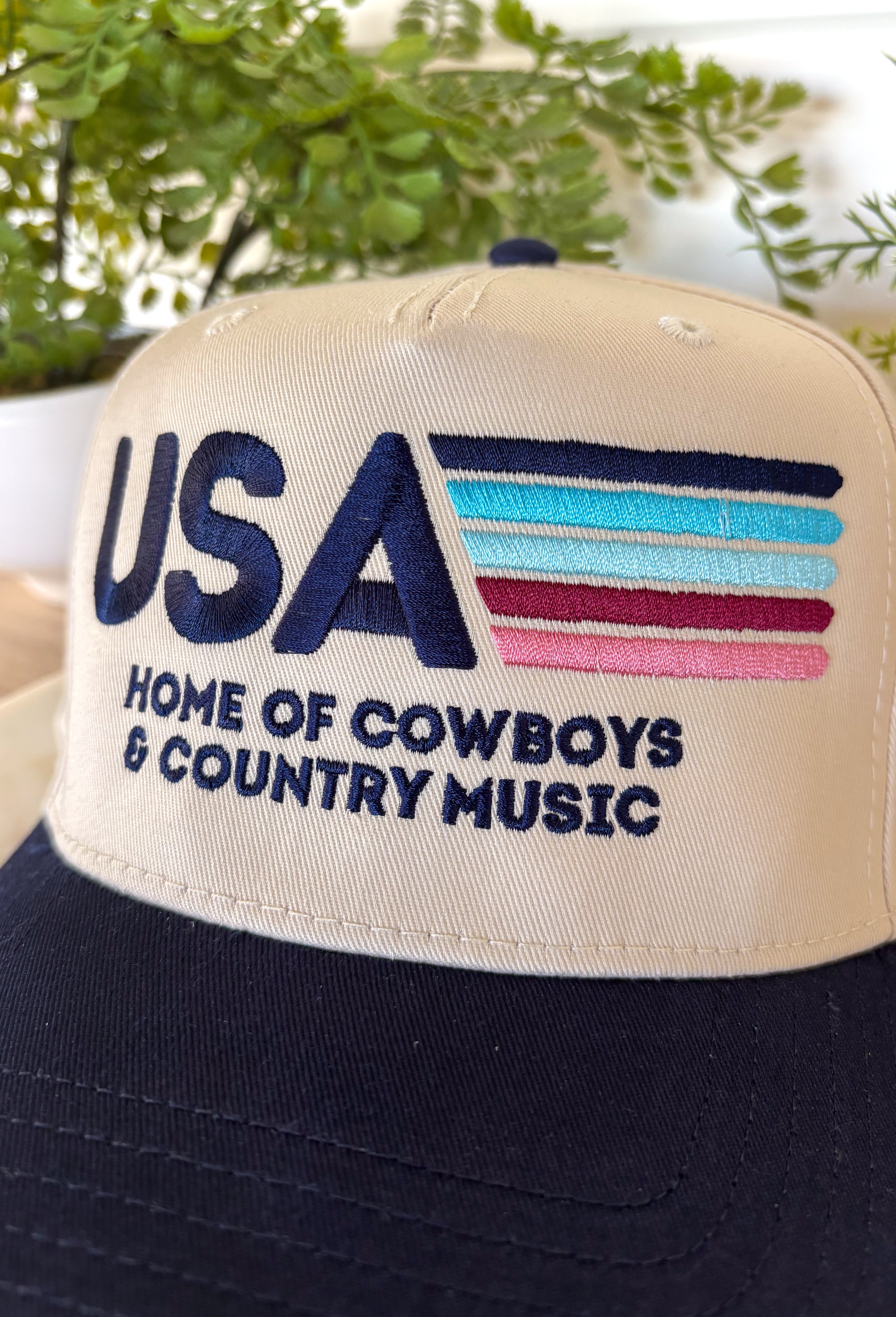 This neutral trucker hat is the perfect laid-back statement for everyday wear. Featuring “USA Home of Cowboys &amp; Country Music” embroidered in navy with colorful stitched accents, it adds a fun pop of detail while keeping things classic and easy to style. The neutral base makes it an effortless match with just about anything, and the adjustable back ensures a comfortable, just-right fit. Whether you’re running errands, heading to a concert, or finishing off a casual outfit, this hat brings a little perso