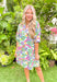 Coastal Views Dress, short puff sleeve v-neck dress with ruffling around the neckline. Palm leaf and floral print covers the white background in the colors amazon green, royal fuchsia, tomato red, lime green, yellow, and blueish purple