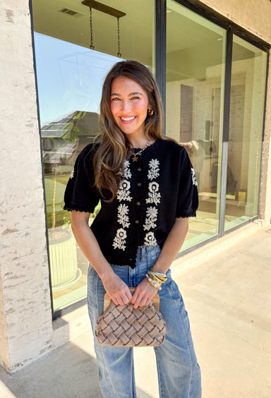 Sweet details and cozy texture make this cardigan a seasonal favorite. Designed in a soft black knit, it’s accented with cream embroidered florals down the front, and on the sleeves for a touch of charm. The scalloped trim along the sleeves, hem, and neckline adds a feminine finish, while the short sleeve and button-down design keep it versatile. An easy layering piece you can style casual or dressed up, this cardigan is the perfect balance of cozy and polished.