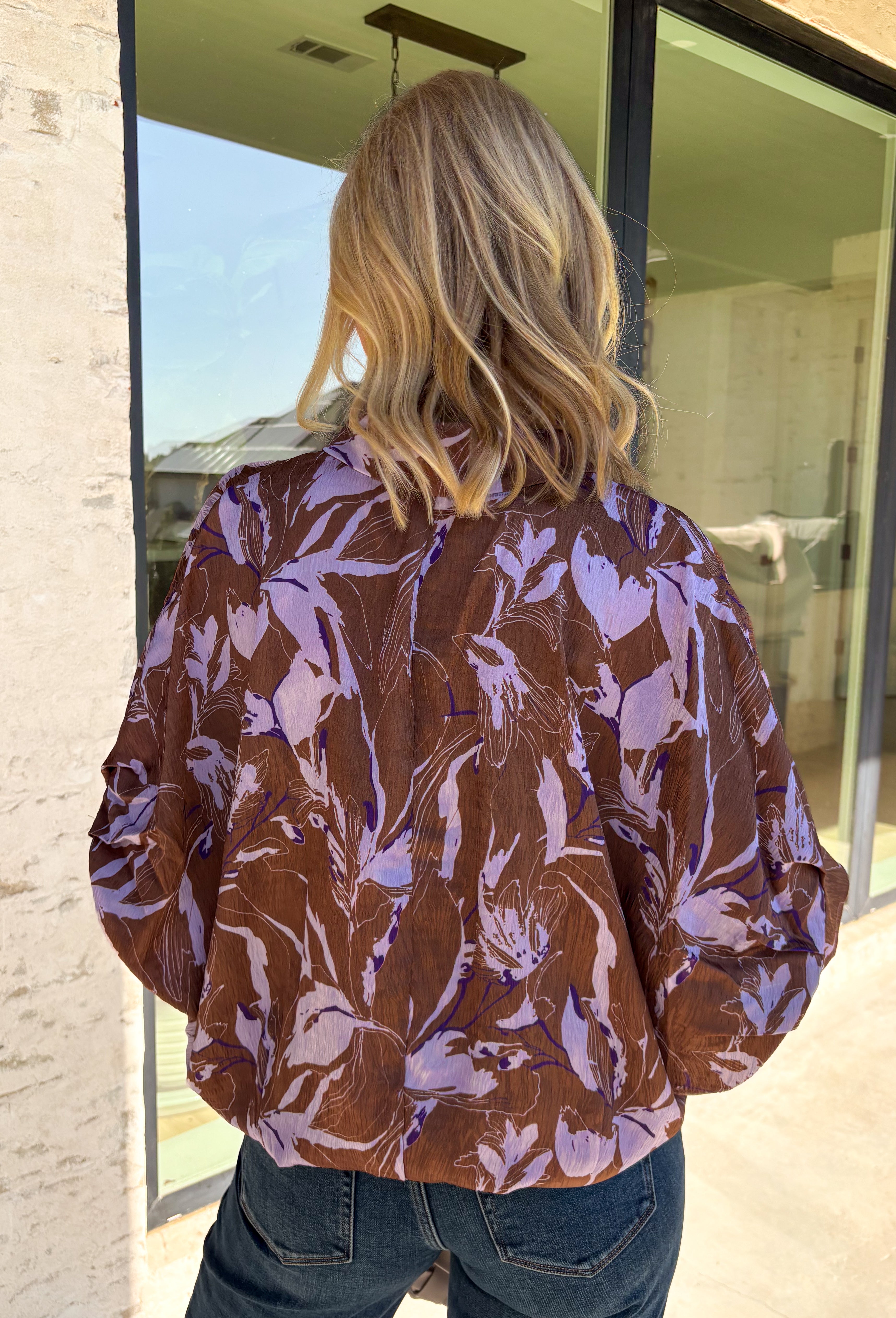 Rich fall shades and standout details make this printed blouse from Entro an easy way to bring a little color into your autumn wardrobe. The warm brown base is accented with lilac and dark purple throughout, creating a unique print that pairs beautifully with the season’s deeper tones. A classic shirt collar and hammered gold buttons dress up the front, while the ¾ dolman sleeves give it a relaxed, comfortable silhouette. 