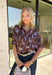 Rich fall shades and standout details make this printed blouse from Entro an easy way to bring a little color into your autumn wardrobe. The warm brown base is accented with lilac and dark purple throughout, creating a unique print that pairs beautifully with the season’s deeper tones. A classic shirt collar and hammered gold buttons dress up the front, while the ¾ dolman sleeves give it a relaxed, comfortable silhouette. 