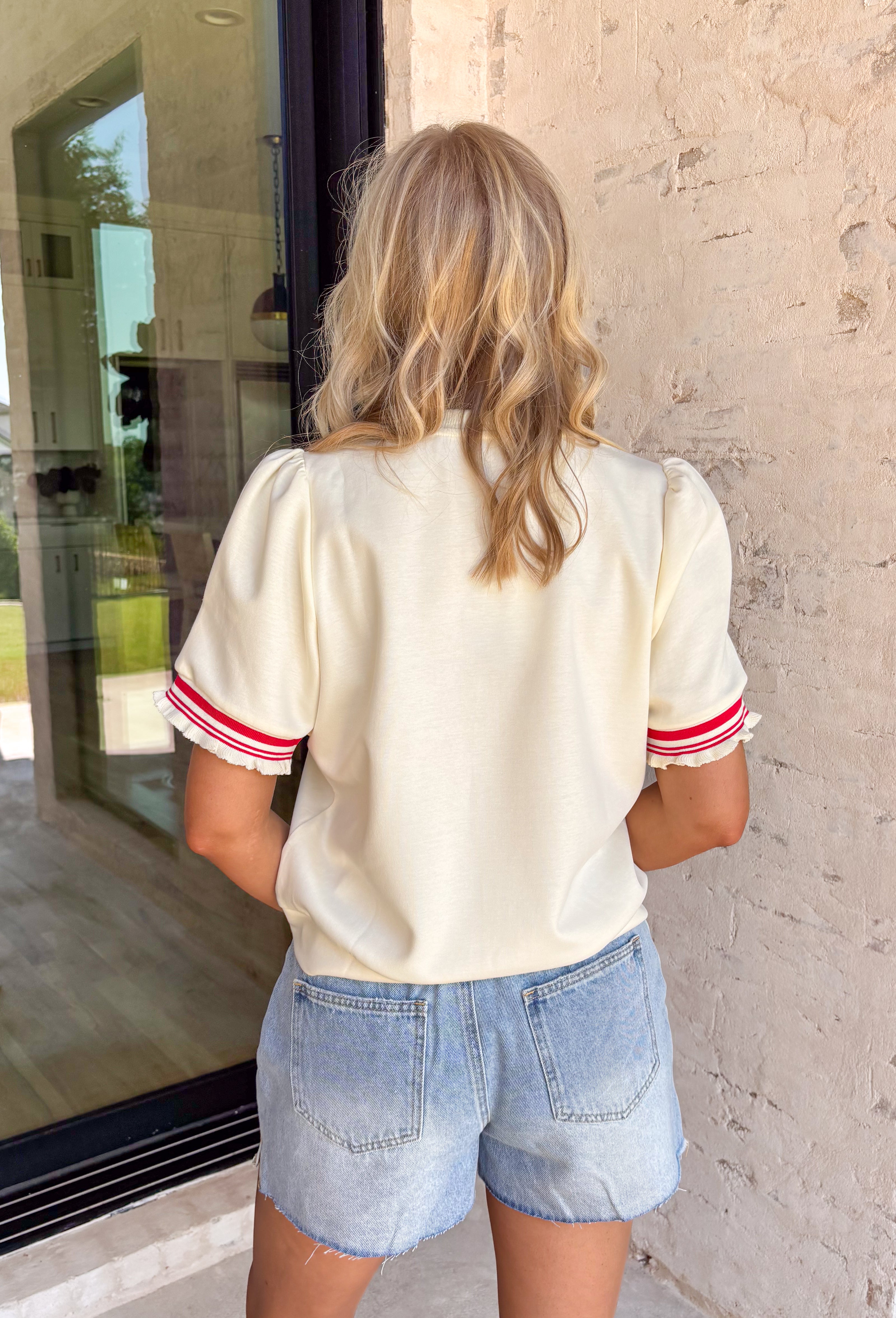This off white short sleeve top is the perfect mix of classic and fun for summer! The clean, neutral base keeps it easy to style, while the red stripe detail on the sleeves adds that sporty pop of color. The front button detail gives it a polished, elevated touch, and the ruffle cuffs bring just the right amount of feminine flair. Lightweight with a soft stretch, it’s comfortable enough for all-day wear—whether you're running errands or meeting up with friends. 