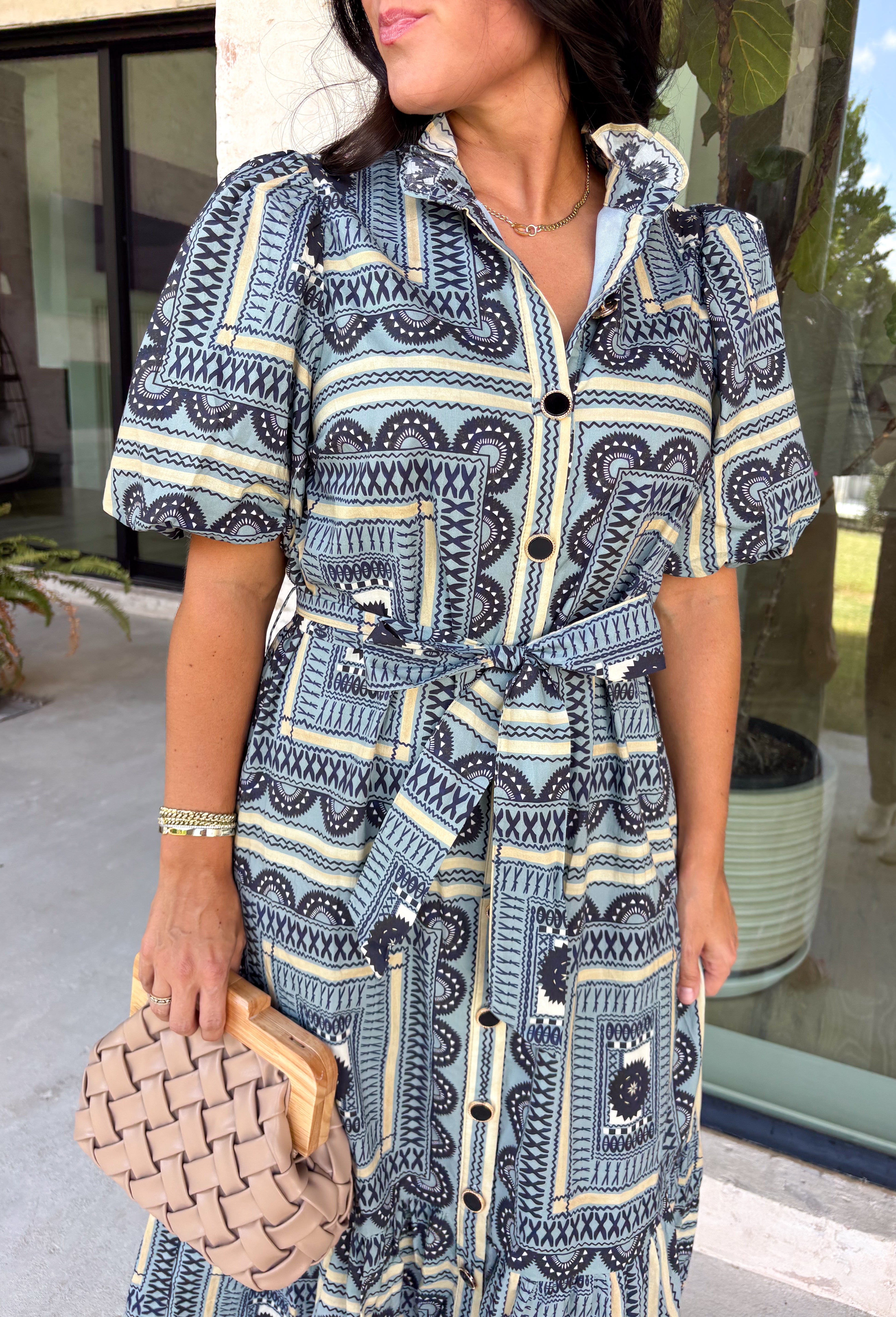 Bring effortless elegance to your wardrobe with this beautifully detailed dress from Entro. Designed with a soft light blue backdrop and an eye-catching abstract print in shades of navy, white, and cream, this timeless piece is perfect for everything from brunch dates to special gatherings. A subtle ruffle neckline adds a feminine touch, while the black buttons outlined in gold create an elevated, polished finish. 