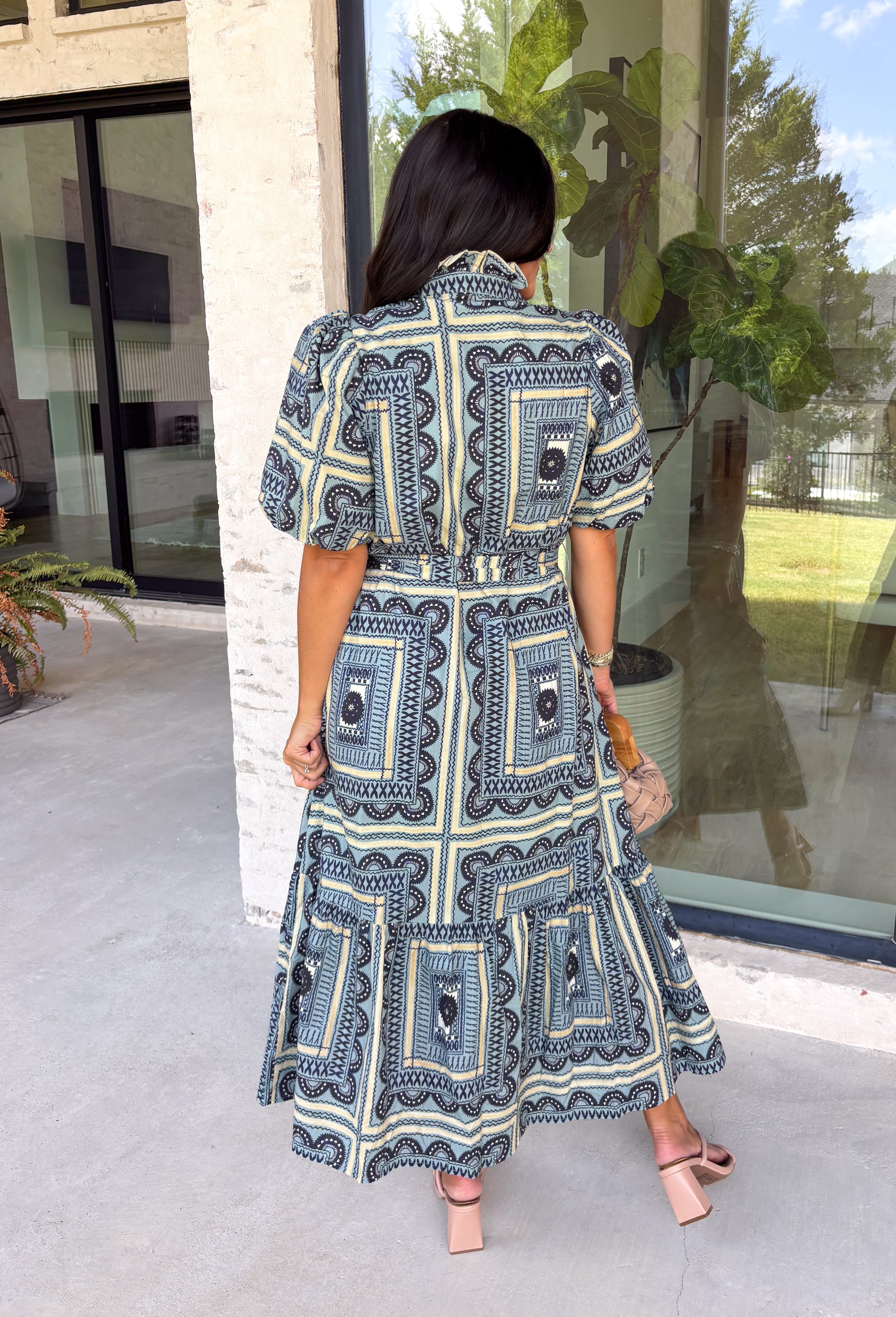 Bring effortless elegance to your wardrobe with this beautifully detailed dress from Entro. Designed with a soft light blue backdrop and an eye-catching abstract print in shades of navy, white, and cream, this timeless piece is perfect for everything from brunch dates to special gatherings. A subtle ruffle neckline adds a feminine touch, while the black buttons outlined in gold create an elevated, polished finish.