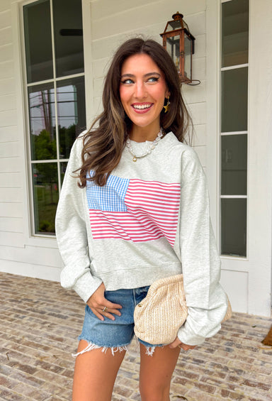 This heather grey pullover is the perfect cozy-meets-festive piece for your summer celebrations! Featuring red and white stripes with a blue and white gingham flag, it brings a fun, playful take on classic patriotic style. The mock neck adds a slightly elevated feel, while the soft cotton fabric keeps it comfortable for those breezy summer nights—think fireworks, lake evenings, or early morning coffee runs. It’s an easy throw-on piece that still feels festive and put together. 