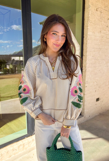 Classic stripes with a spring twist—this tan and off-white striped top by Entro is full of charm and elevated detail. Featuring a band collar v-neck with contrast binding, it has a polished look that’s still easy and effortless. The 3/4 puff sleeves steal the show with colorful floral embroidery in shades of pink, peach, and green, adding the prettiest pop of color and texture. Finished with button cuffs, this top is perfect for dressing up denim or adding a little something special to your everyday spring 