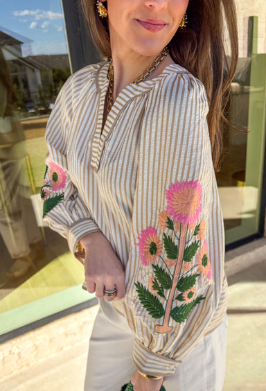 Classic stripes with a spring twist—this tan and off-white striped top by Entro is full of charm and elevated detail. Featuring a band collar v-neck with contrast binding, it has a polished look that’s still easy and effortless. The 3/4 puff sleeves steal the show with colorful floral embroidery in shades of pink, peach, and green, adding the prettiest pop of color and texture. Finished with button cuffs, this top is perfect for dressing up denim or adding a little something special to your everyday spring 