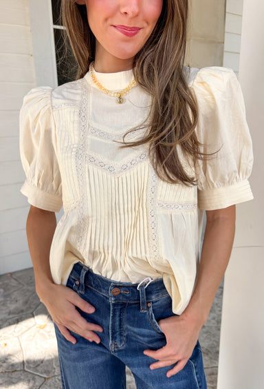 Soft, feminine, and effortlessly timeless, this beautiful ecru top is the kind of piece you'll treasure season after season! Crafted from breathable cotton, it features delicate lace detailing and chic pleating throughout for a look that's both romantic and refined. The puff sleeves add just the right amount of charm, while the keyhole button closure at the back provides a graceful finishing touch. 