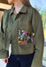 Make a statement everywhere you go with this stunning denim jacket from Veveret. Crafted in a rich olive denim, this standout layer features a breathtaking sheer embroidered floral design across the back, accented with shimmering sequins in shades of green, pink, orange, and brown. Coordinating floral sequin details on the front pockets tie the look together beautifully, while the classic collared neckline and button-front design keep it timeless. 