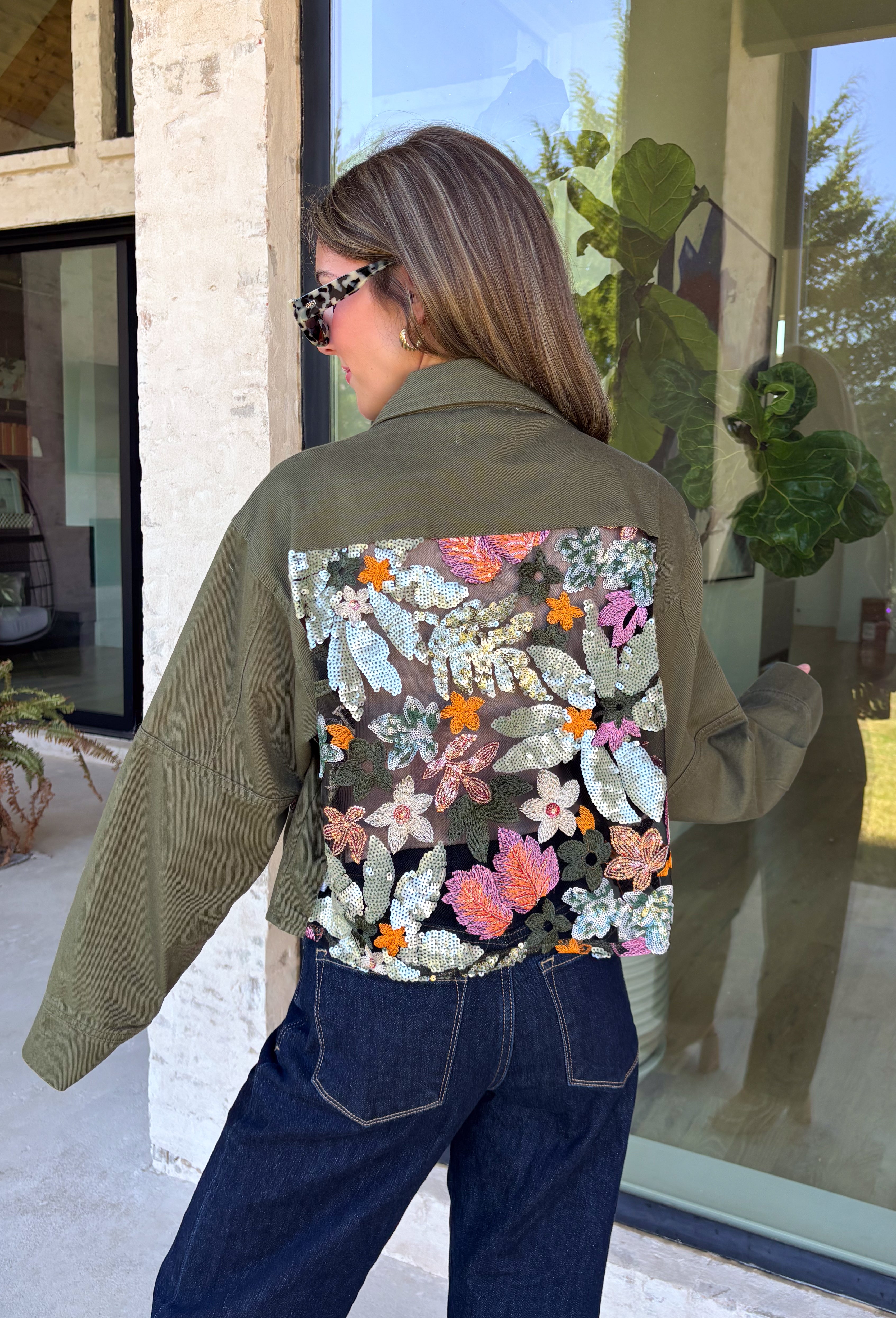 Make a statement everywhere you go with this stunning denim jacket from Veveret. Crafted in a rich olive denim, this standout layer features a breathtaking sheer embroidered floral design across the back, accented with shimmering sequins in shades of green, pink, orange, and brown. Coordinating floral sequin details on the front pockets tie the look together beautifully, while the classic collared neckline and button-front design keep it timeless. 