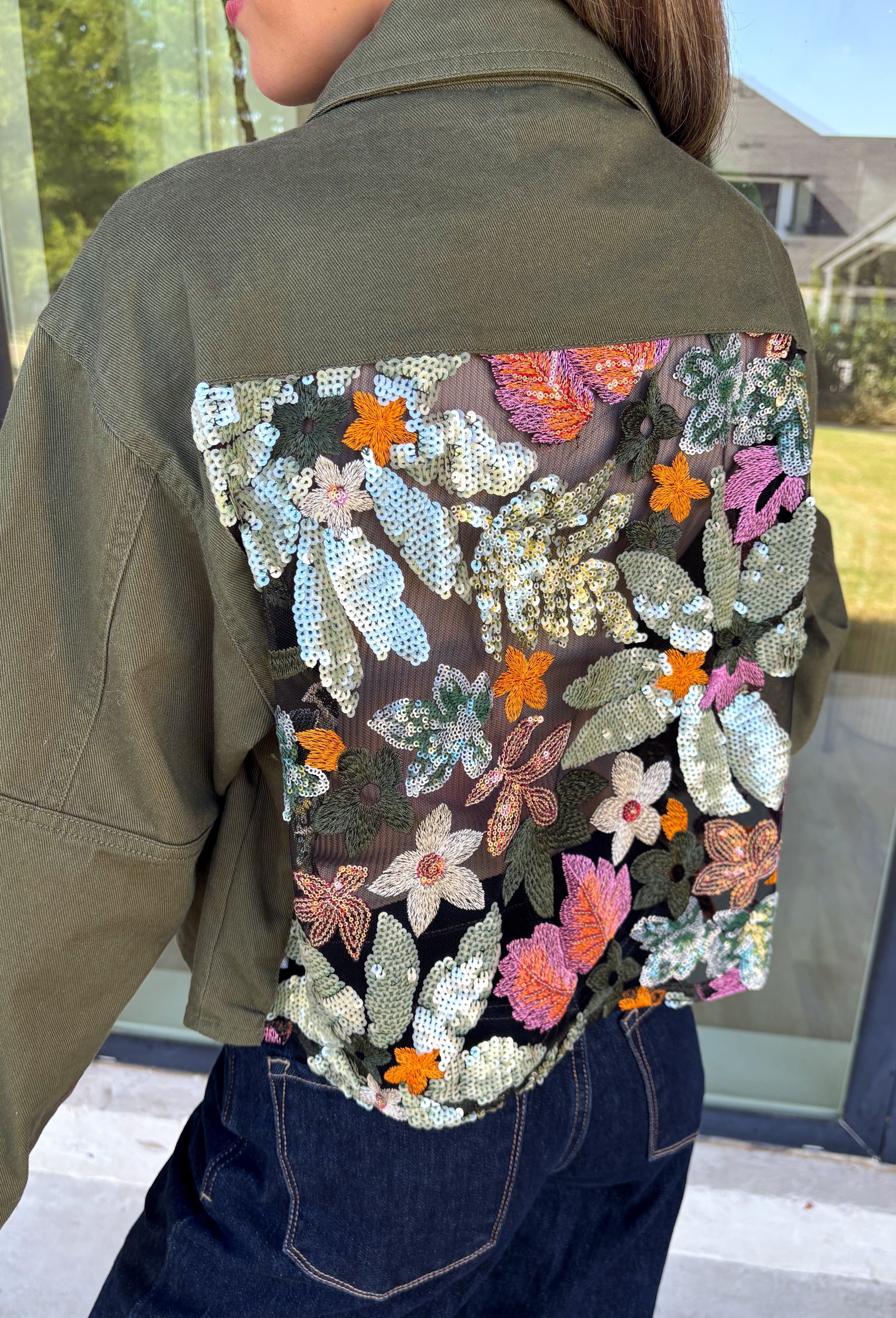 Make a statement everywhere you go with this stunning denim jacket from Veveret. Crafted in a rich olive denim, this standout layer features a breathtaking sheer embroidered floral design across the back, accented with shimmering sequins in shades of green, pink, orange, and brown. Coordinating floral sequin details on the front pockets tie the look together beautifully, while the classic collared neckline and button-front design keep it timeless. 