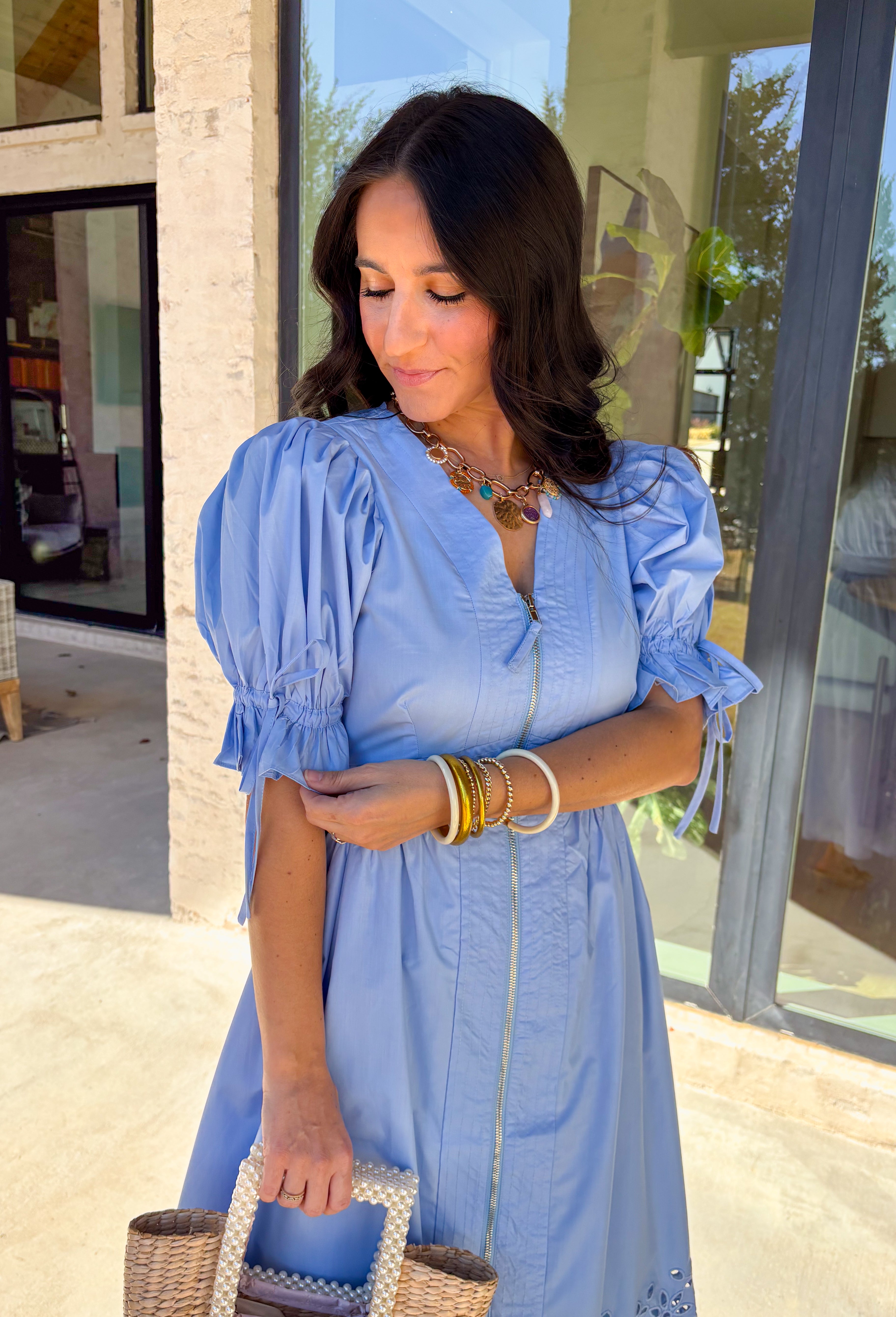 A statement piece with a touch of whimsy, this light blue midi dress is designed to turn heads. Short puff sleeves and a flattering stitch-accent v-neckline pair beautifully with a defined waistband, while the embroidered skirt hem adds that extra bit of charm. The zipper front closure offers a modern twist, and the smocked waistband at the back ensures all-day comfort. This is perfect for any spring occasion!