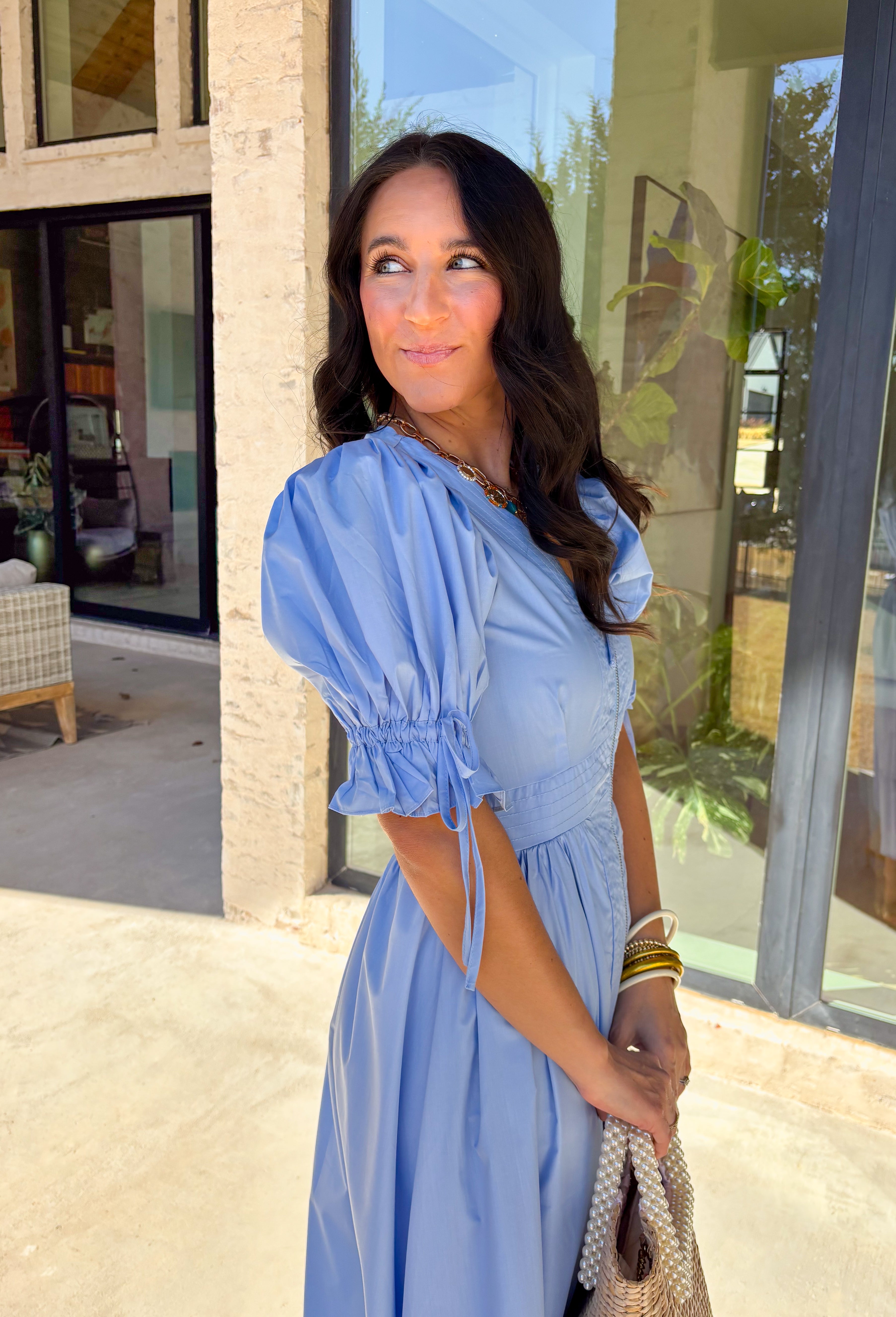 A statement piece with a touch of whimsy, this light blue midi dress is designed to turn heads. Short puff sleeves and a flattering stitch-accent v-neckline pair beautifully with a defined waistband, while the embroidered skirt hem adds that extra bit of charm. The zipper front closure offers a modern twist, and the smocked waistband at the back ensures all-day comfort. This is perfect for any spring occasion!