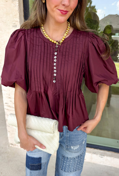 Elevate your everyday wardrobe with this beautifully detailed top from Entro. Designed in a rich merlot hue, this timeless blouse features elegant pleating throughout for added texture and a polished finish. Decorative front button details add classic charm, while the puff sleeves create a feminine silhouette that's easy to dress up or down. Crafted from a lightweight fabric blend, this versatile top is perfect for everything from workdays to weekend brunches and seasonal gatherings.