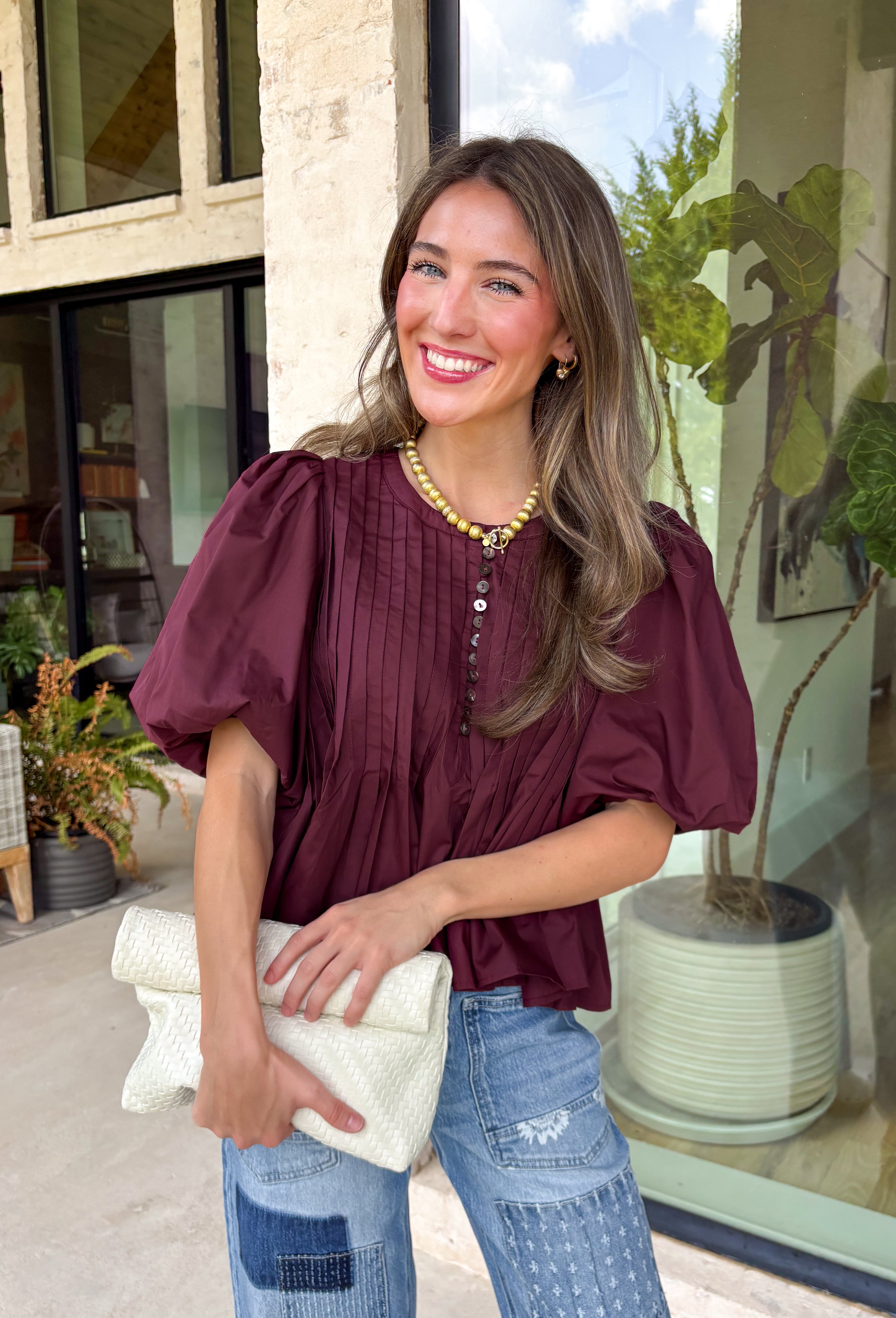 Elevate your everyday wardrobe with this beautifully detailed top from Entro. Designed in a rich merlot hue, this timeless blouse features elegant pleating throughout for added texture and a polished finish. Decorative front button details add classic charm, while the puff sleeves create a feminine silhouette that's easy to dress up or down. Crafted from a lightweight fabric blend, this versatile top is perfect for everything from workdays to weekend brunches and seasonal gatherings.