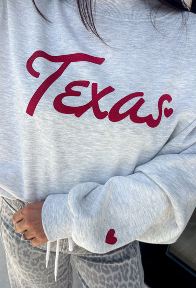 Show your Texas pride in the coziest way possible! This ash grey mock neck sweatshirt blends everyday comfort with timeless style, featuring "Texas" in soft script across the chest and a sweet heart detail on the sleeve. The fleece-lined interior keeps you warm and cozy, while the classic silhouette makes it an easy favorite for everything from football Saturdays and coffee runs to relaxed weekends at home. 