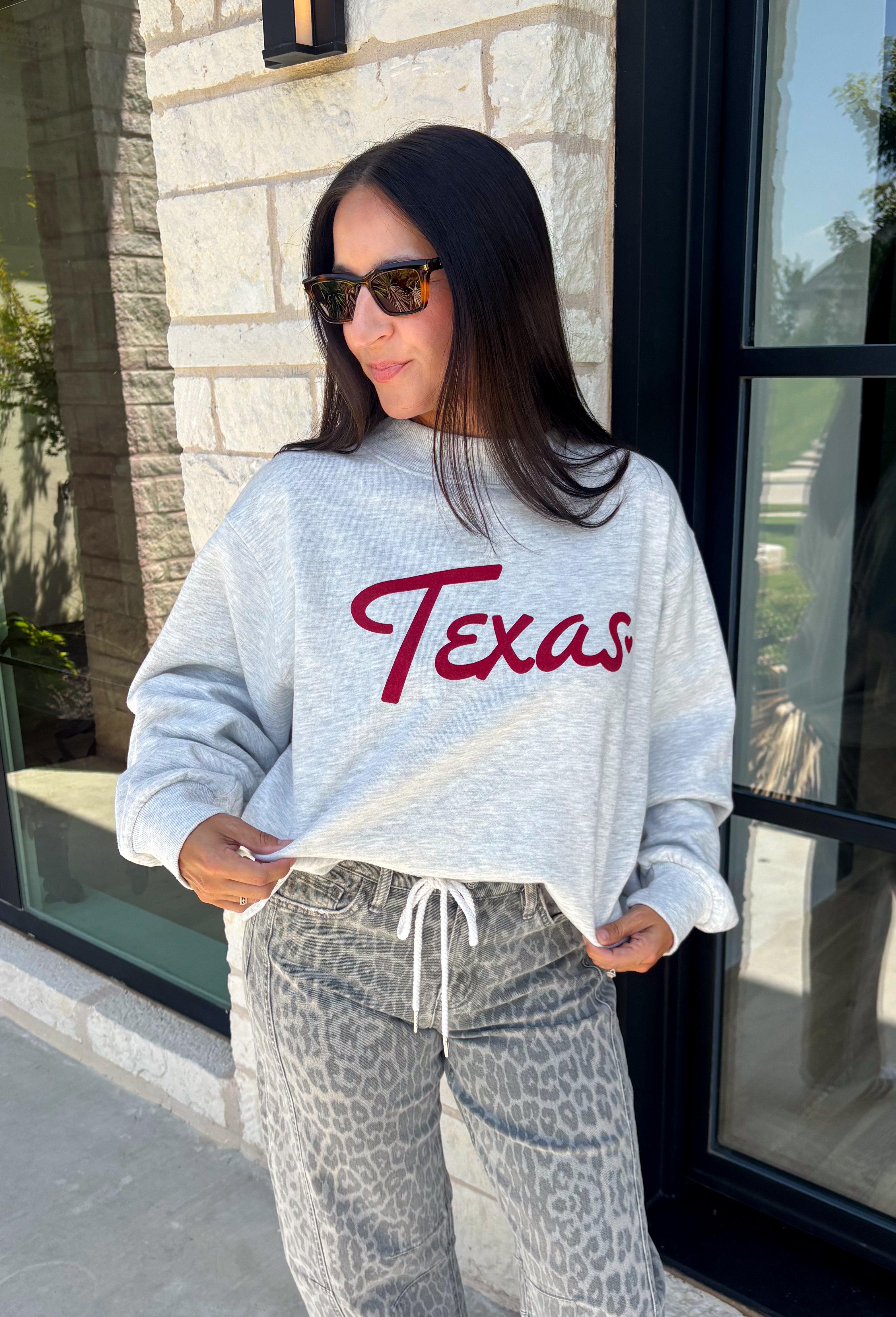 Show your Texas pride in the coziest way possible! This ash grey mock neck sweatshirt blends everyday comfort with timeless style, featuring "Texas" in soft script across the chest and a sweet heart detail on the sleeve. The fleece-lined interior keeps you warm and cozy, while the classic silhouette makes it an easy favorite for everything from football Saturdays and coffee runs to relaxed weekends at home. 