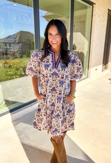 Romantic florals meet cozy charm in this cream short sleeve dress. Designed with shades of burgundy and blue for an autumn-inspired palette, it features a botanical print and contrast binding along the v-neckline for a polished finish. The puff sleeves add a playful touch, while the ruffle hem brings soft movement. Fully lined with side pockets for practicality, this dress is as easy to wear as it is beautiful.
