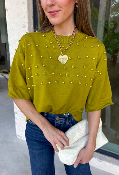 A rich olive green gets a pretty update with this pearl-detailed short sleeve sweater from Glam. Pearl detailing throughout adds a special touch to the soft knit, making it easy to dress up while still keeping that cozy, everyday feel. Exposed hems bring in a relaxed finish, while ribbed detailing along the neckline, sleeve cuffs, and hem gives it a classic sweater look. Wear it to the office, dinner plans, or anytime your outfit could use a little something extra.