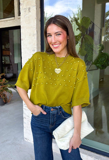 A rich olive green gets a pretty update with this pearl-detailed short sleeve sweater from Glam. Pearl detailing throughout adds a special touch to the soft knit, making it easy to dress up while still keeping that cozy, everyday feel. Exposed hems bring in a relaxed finish, while ribbed detailing along the neckline, sleeve cuffs, and hem gives it a classic sweater look. Wear it to the office, dinner plans, or anytime your outfit could use a little something extra.