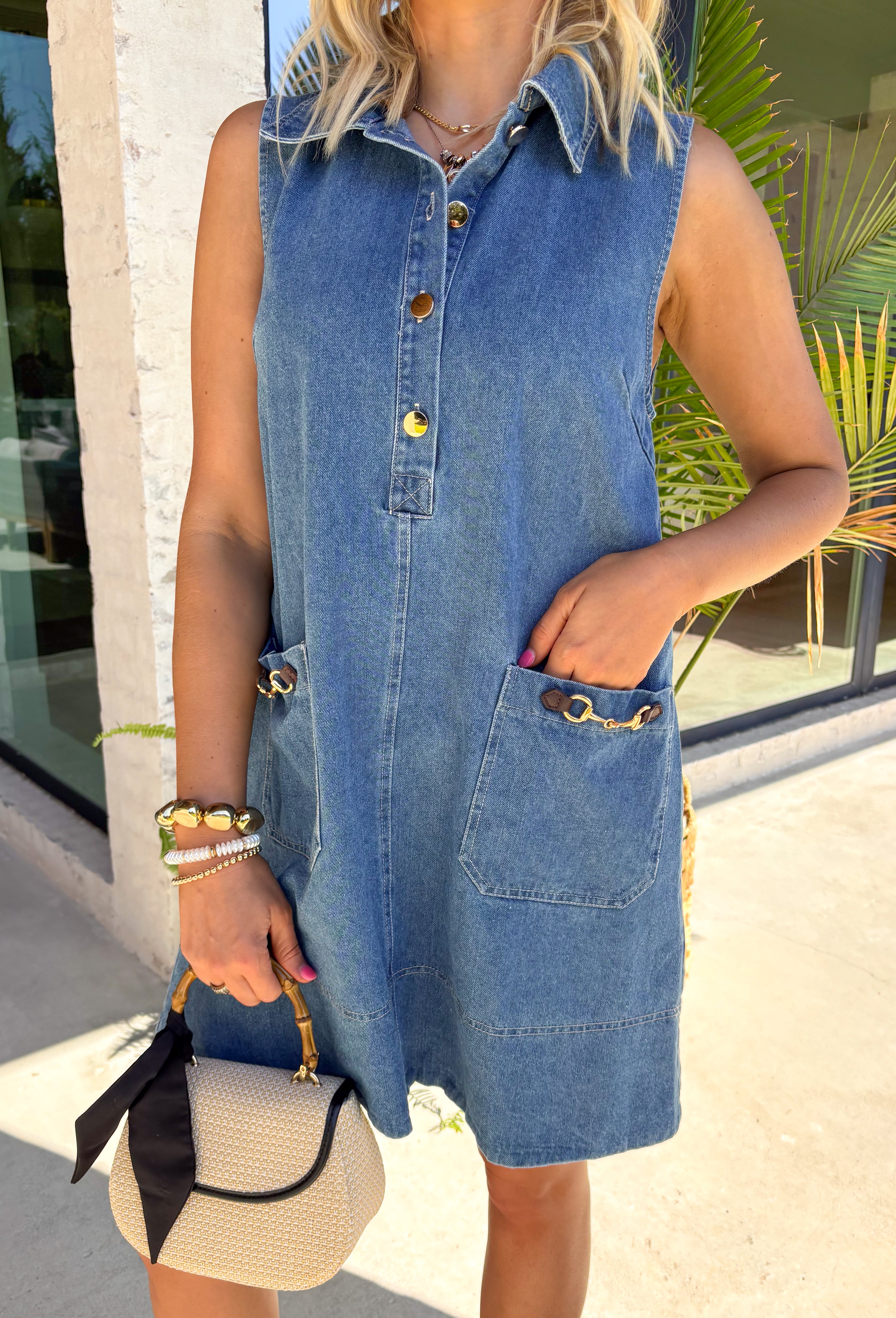 This sleeveless denim mini dress is the perfect mix of classic and cool for summer. Designed with a button front and front flap pockets featuring buckle accents, it brings a subtle utility vibe that feels both trendy and timeless. The lightweight cotton keeps it breathable for warm days, while the structured shape makes it easy to dress up or down. Whether you're heading to brunch, running errands, or packing for vacation, this is one of those throw-on-and-go pieces that always works. 
