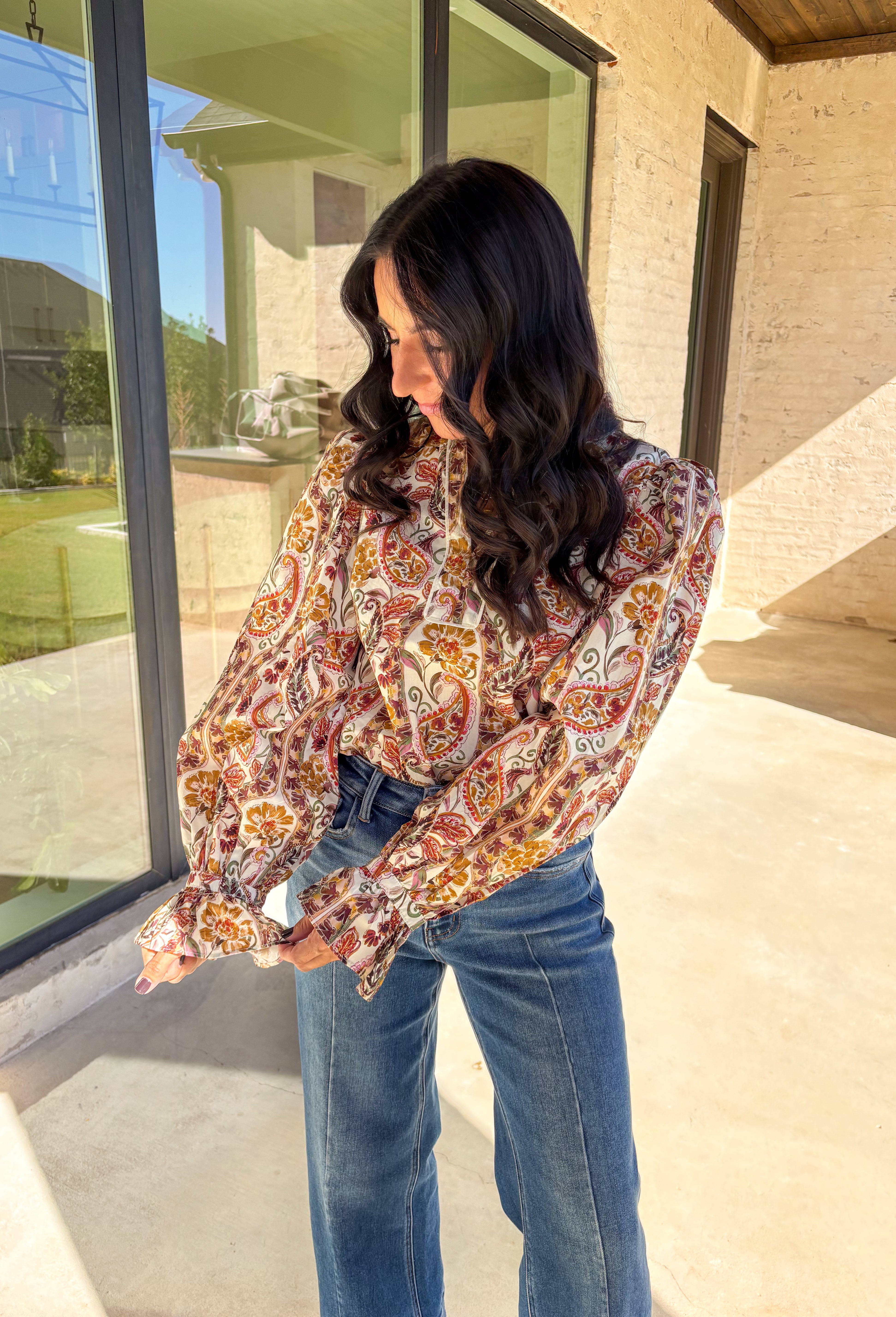 Effortlessly feminine and fall-ready, this neutral paisley print top by Entro brings timeless charm to your wardrobe. Designed with a ruffled band collar and a v-neckline, it offers just the right mix of structure and softness. The buttoned cuffs with delicate ruffle edges add a romantic finishing touch, making it easy to dress up or wear casually through the season.