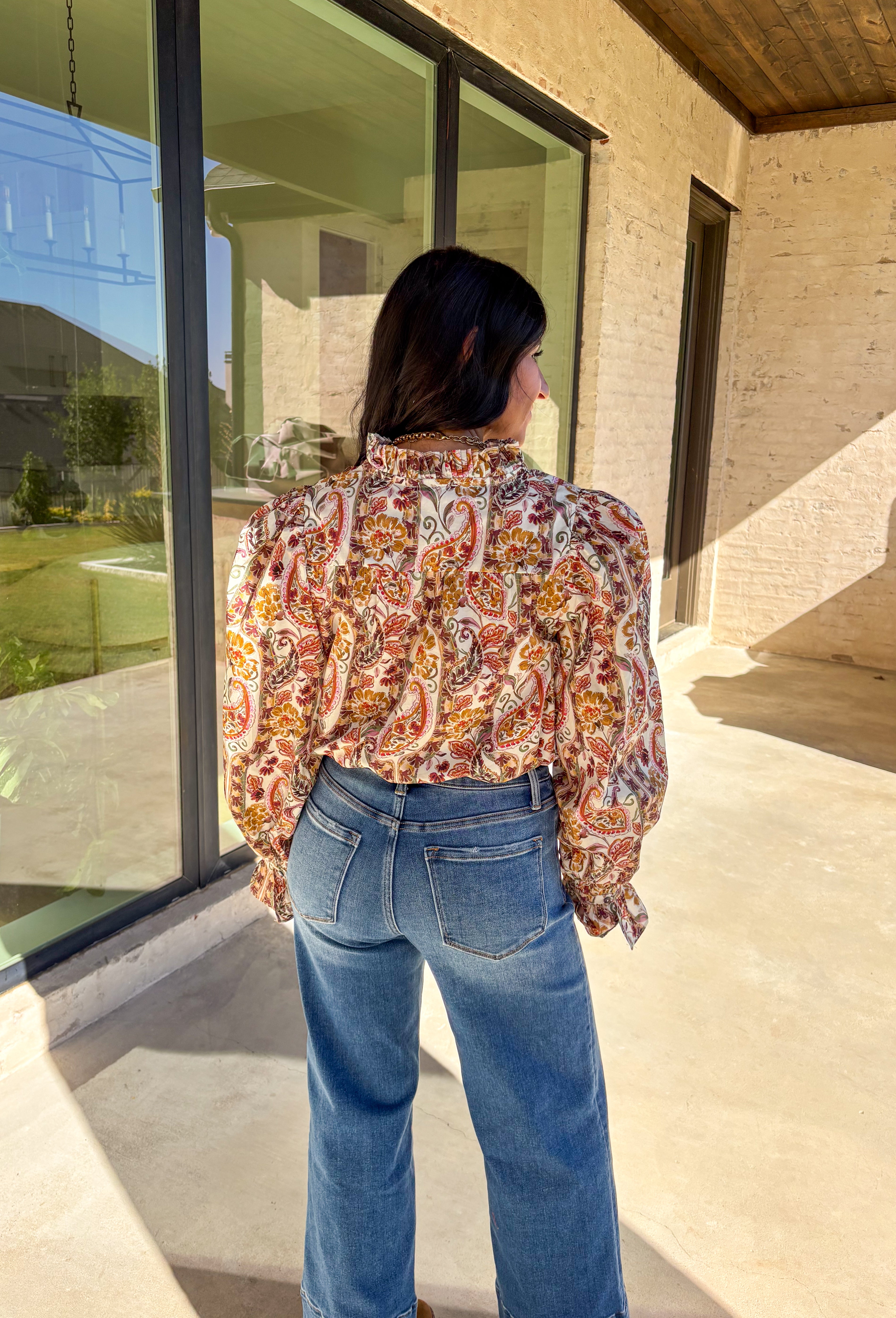 Effortlessly feminine and fall-ready, this neutral paisley print top by Entro brings timeless charm to your wardrobe. Designed with a ruffled band collar and a v-neckline, it offers just the right mix of structure and softness. The buttoned cuffs with delicate ruffle edges add a romantic finishing touch, making it easy to dress up or wear casually through the season.