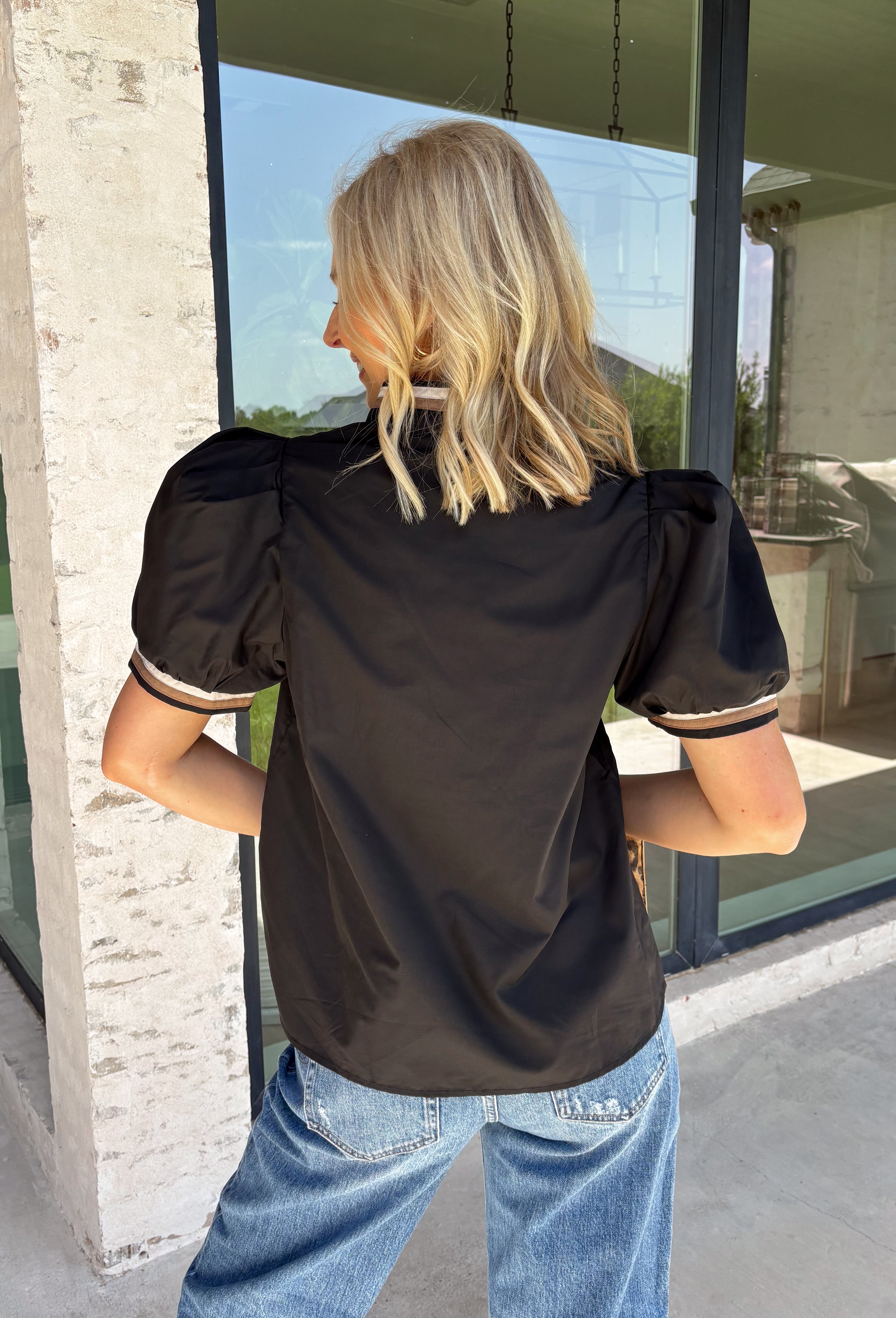 Elevate your everyday wardrobe with this effortlessly polished top from Entro. Designed in a classic black hue, this versatile blouse features beautiful tan and cream contrast binding along the flattering v-neckline for a sophisticated touch. The nonfunctional button detail at the front adds timeless charm, while the short puff sleeves with contrast cuffs create a feminine silhouette. Easy to dress up or down, this top is perfect for workdays, dinner dates, and everything in between.