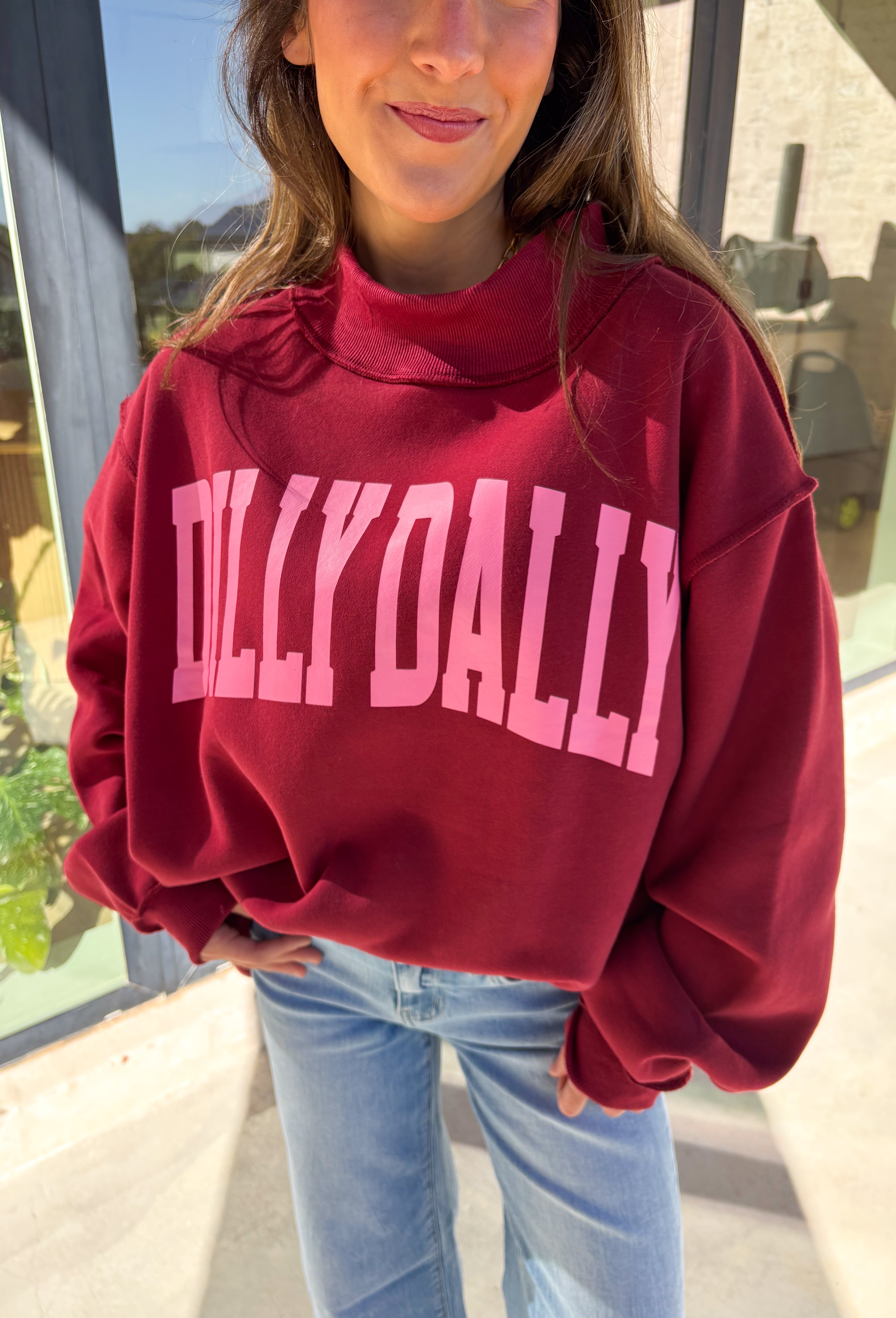 Make your winter wardrobe a little more fun with this playful “Dilly Dally” pullover by Bucket List. Featuring bold hot pink lettering on a rich burgundy backdrop, this cozy mock neck sweatshirt is made for chill days and cheerful moods. With its oversized fit and soft fabric, it’s the perfect mix of comfort and attitude for the season.