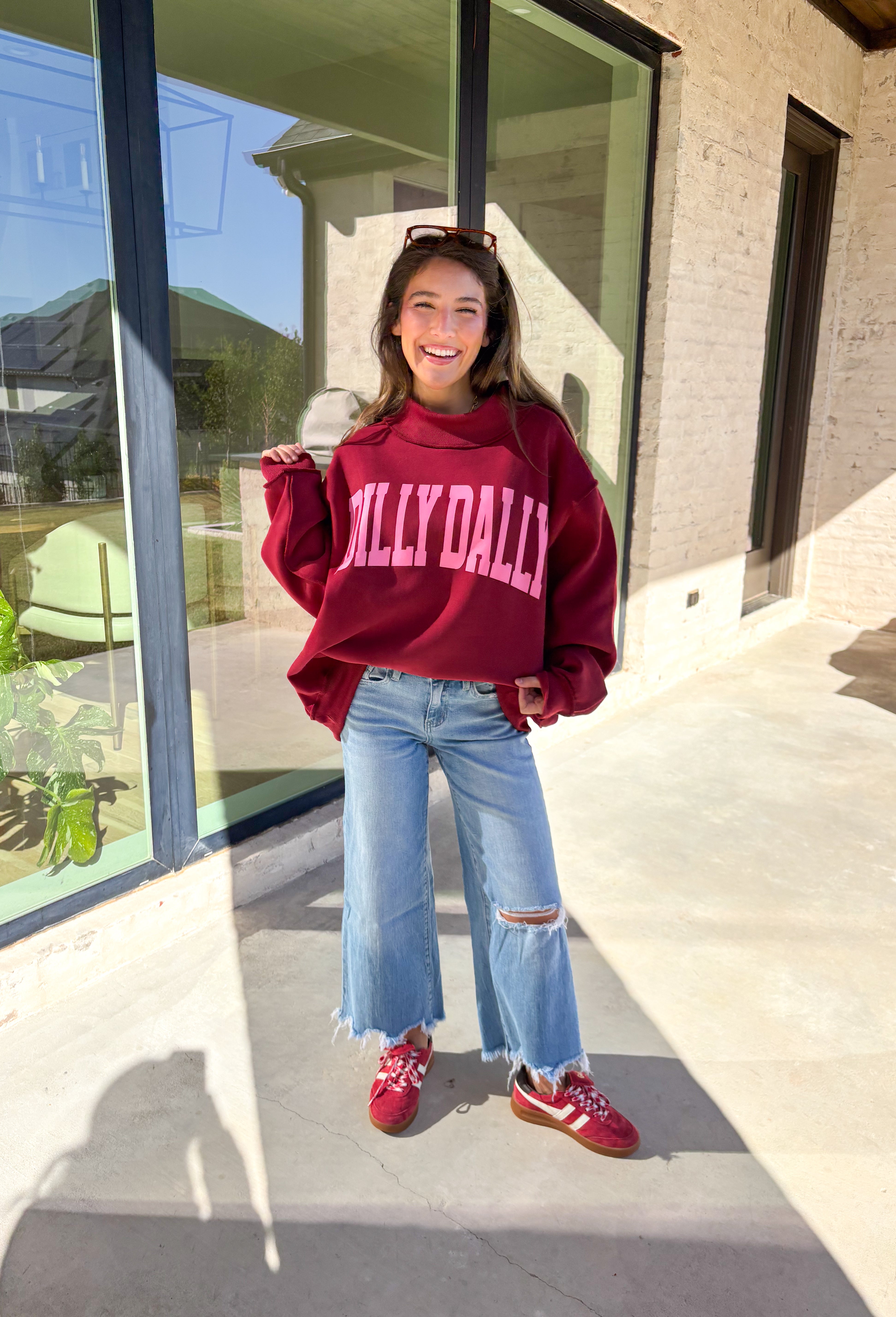 Make your winter wardrobe a little more fun with this playful “Dilly Dally” pullover by Bucket List. Featuring bold hot pink lettering on a rich burgundy backdrop, this cozy mock neck sweatshirt is made for chill days and cheerful moods. With its oversized fit and soft fabric, it’s the perfect mix of comfort and attitude for the season.