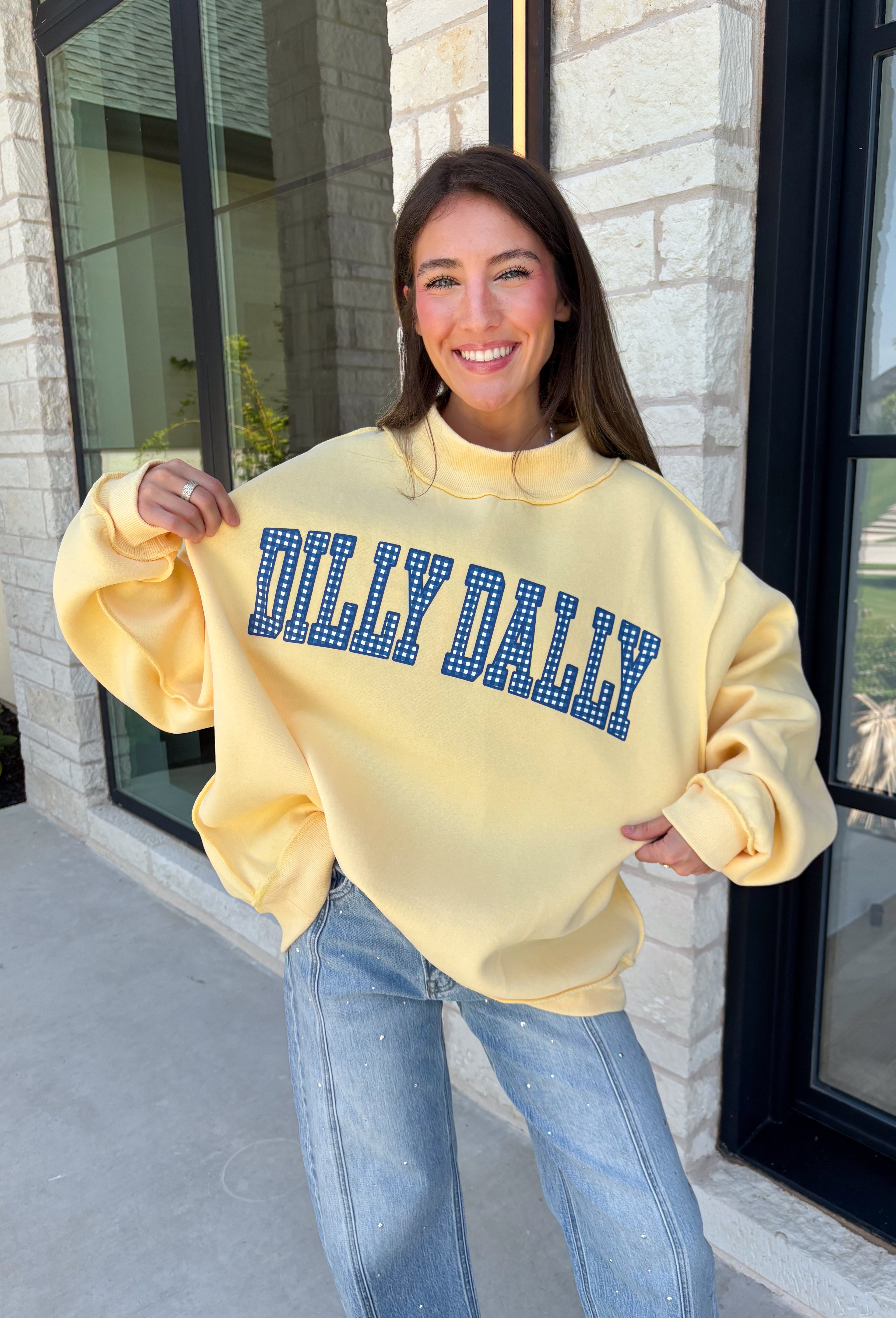 Why choose one look when you can have two? This cozy reversible pullover is full of personality with playful sayings on each side. One side features "Dilly Dally" in blue and white gingham lettering, while the fleece-lined reverse showcases "Lolly Gag" in pink and white gingham for an equally fun look. Designed with an oversized fit and a classic mock neckline, this butter yellow sweatshirt is perfect for slow mornings, coffee runs, travel days, and all your cozy fall adventures.