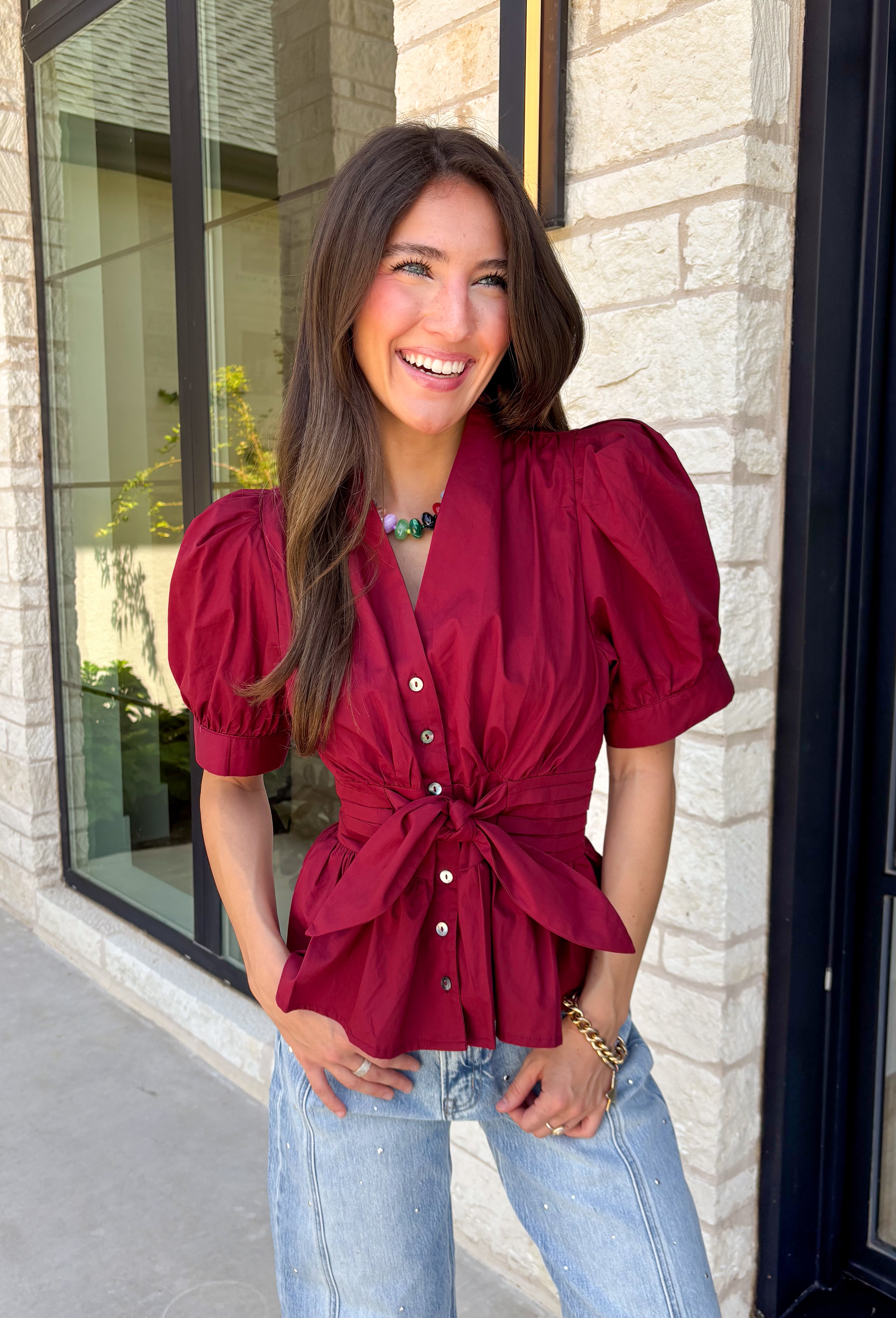 Rich seasonal color and flattering details make this merlot blouse a must-have for fall! Crafted from breathable cotton, this feminine top features a button front, soft puff sleeves, and a flattering tie waist that beautifully defines your silhouette. Pleating at the waist adds a polished touch, while the smocked back provides all-day comfort and the perfect fit. 