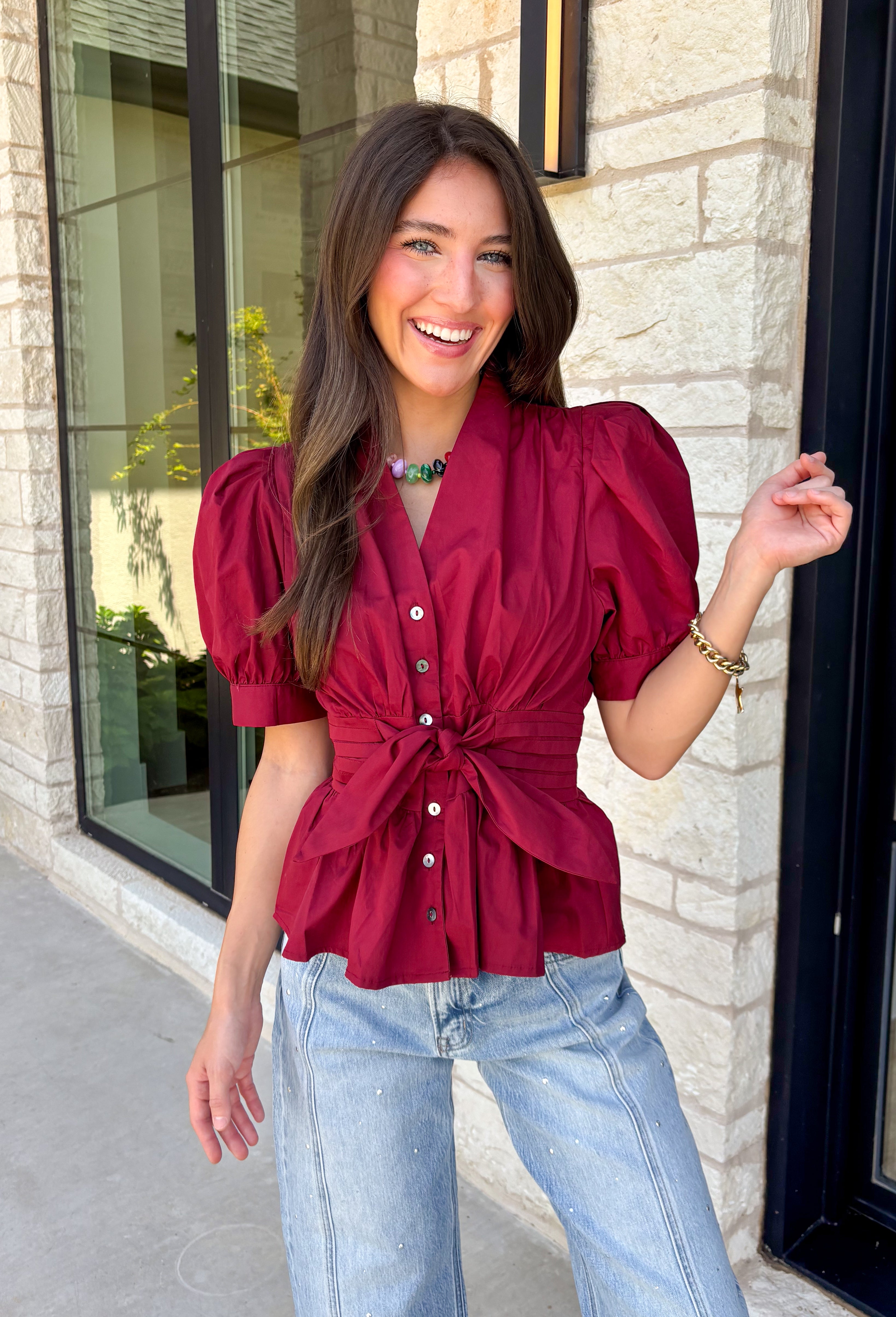 Rich seasonal color and flattering details make this merlot blouse a must-have for fall! Crafted from breathable cotton, this feminine top features a button front, soft puff sleeves, and a flattering tie waist that beautifully defines your silhouette. Pleating at the waist adds a polished touch, while the smocked back provides all-day comfort and the perfect fit. 