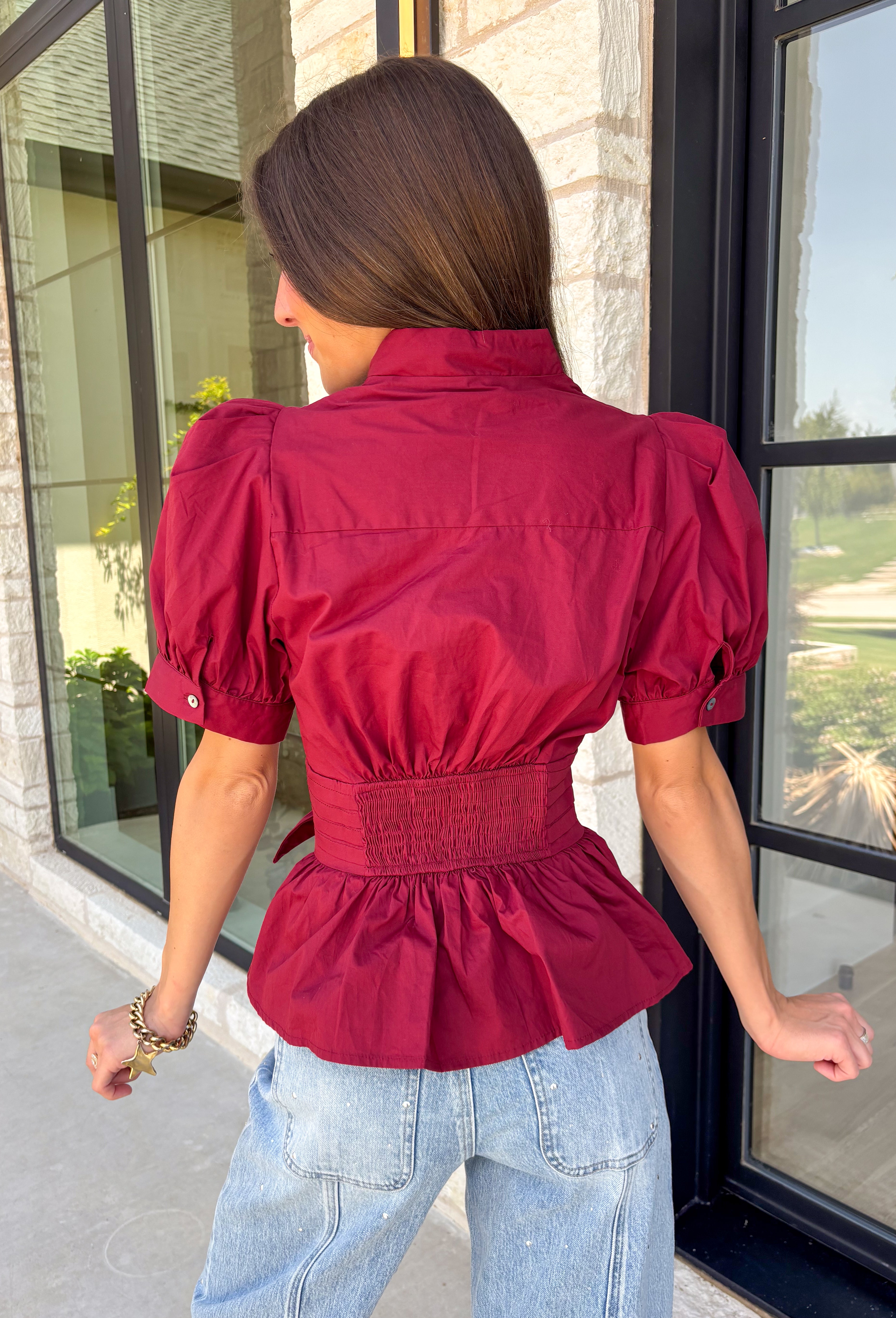 Rich seasonal color and flattering details make this merlot blouse a must-have for fall! Crafted from breathable cotton, this feminine top features a button front, soft puff sleeves, and a flattering tie waist that beautifully defines your silhouette. Pleating at the waist adds a polished touch, while the smocked back provides all-day comfort and the perfect fit. 