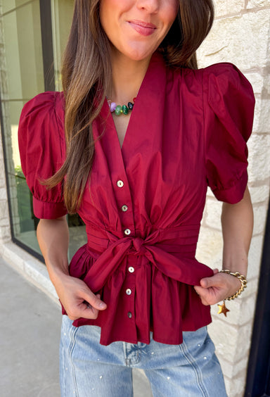 Rich seasonal color and flattering details make this merlot blouse a must-have for fall! Crafted from breathable cotton, this feminine top features a button front, soft puff sleeves, and a flattering tie waist that beautifully defines your silhouette. Pleating at the waist adds a polished touch, while the smocked back provides all-day comfort and the perfect fit. 