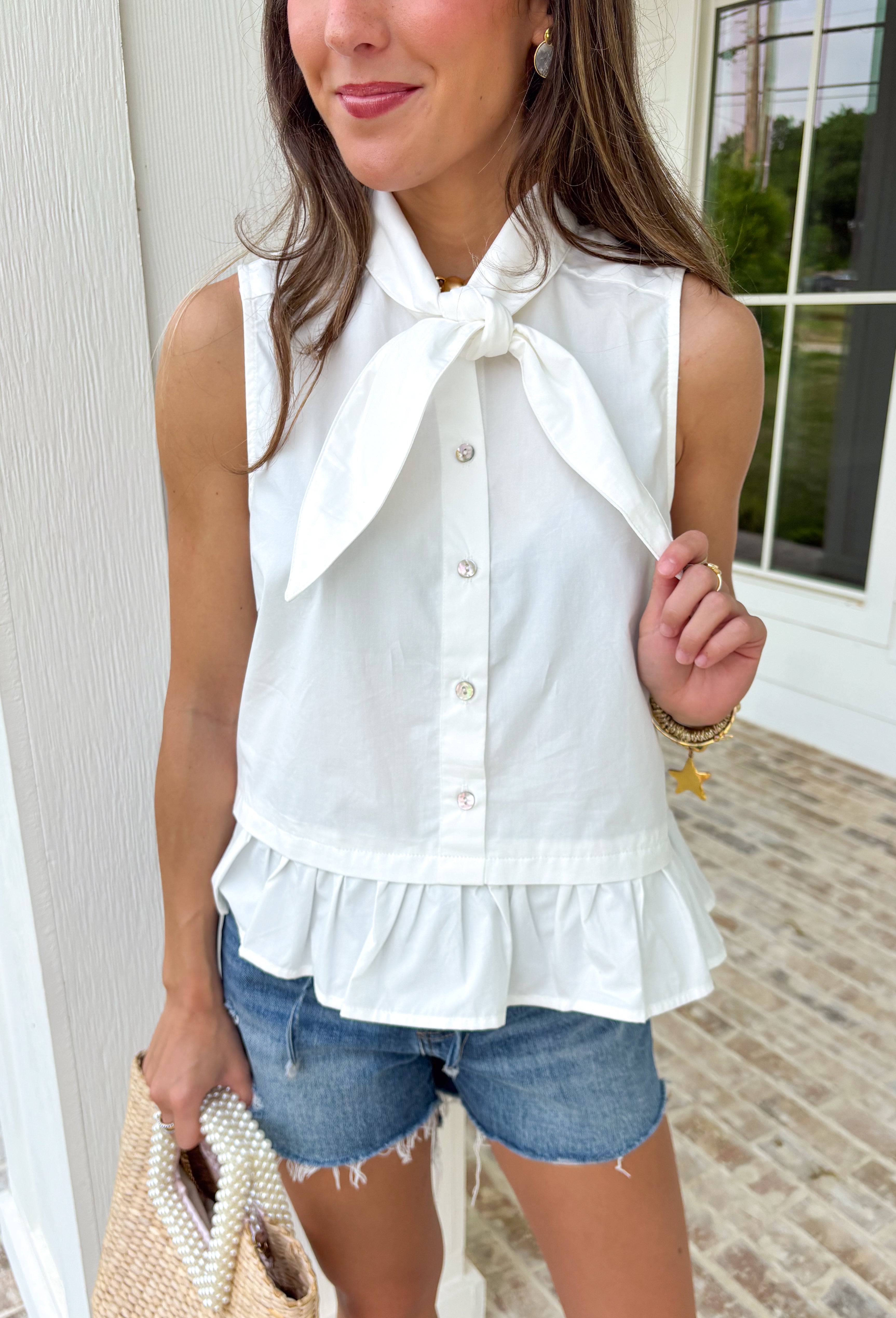 Crisp, classic, and endlessly versatile — this white sleeveless button down top is one you’ll reach for again and again. The clean silhouette keeps it timeless, while the tie detail at the top adds a soft, feminine touch that sets it apart from your basic button downs. Finished with a subtle ruffle hem for a hint of movement and charm, this top is lightweight and breathable in 100% cotton. It’s polished enough for work and easy enough for weekend wear — the kind of staple that works all season long.