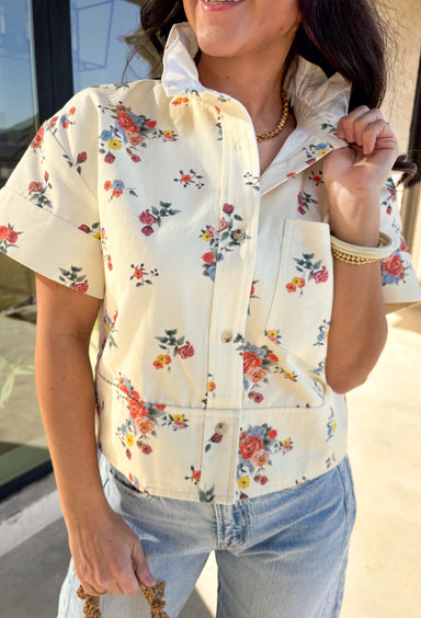 Easy, playful, and perfect for spring, this cream short sleeve button-down top by Entro has that effortless denim-inspired feel with a fun seasonal twist. Designed with a colorful floral print throughout, it brings the prettiest pops of color to a soft, structured cotton base. The ruffle neckline adds a sweet, feminine touch, while the chest pocket keeps the look casual and everyday-ready. This is one of those throw-on tops that instantly makes your outfit feel put together.