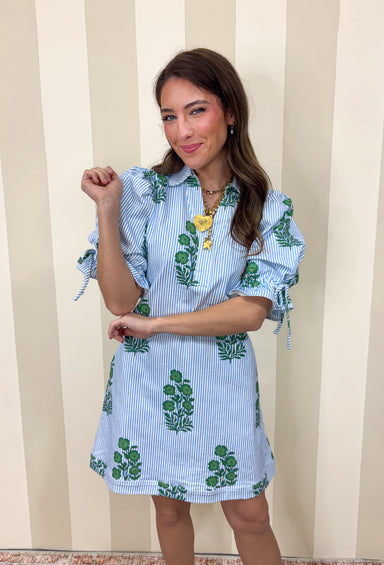 Classic with a cheerful spring twist, this blue and white striped dress by Entro is effortlessly charming. Featuring a crisp stripe base with a green floral print throughout, it feels fresh and timeless all at once. The collared neckline adds a polished touch, while the elastic sleeves with adjustable tie details bring in a playful, feminine finish. Made from a comfortable cotton blend with a hint of stretch, this dress is easy to wear and perfect for sunny days ahead.