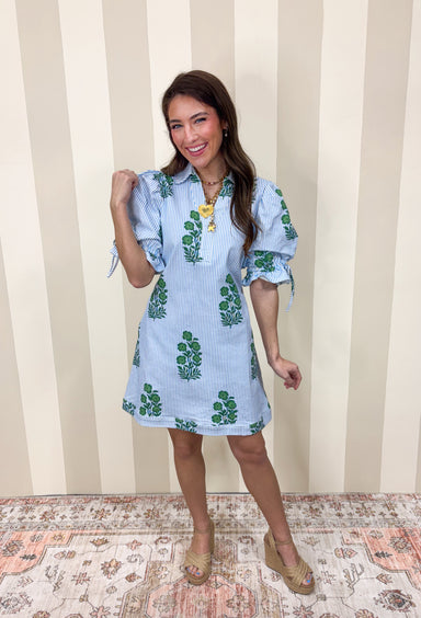 Classic with a cheerful spring twist, this blue and white striped dress by Entro is effortlessly charming. Featuring a crisp stripe base with a green floral print throughout, it feels fresh and timeless all at once. The collared neckline adds a polished touch, while the elastic sleeves with adjustable tie details bring in a playful, feminine finish. Made from a comfortable cotton blend with a hint of stretch, this dress is easy to wear and perfect for sunny days ahead.