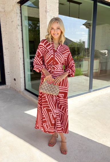 Bring effortless elegance to your fall wardrobe with this beautifully patterned midi dress from Entro. Featuring a soft light pink backdrop with a rich brown geometric print, this timeless dress offers a unique blend of feminine charm and modern style. The flattering v-neckline and removable matching belt create a customizable silhouette, while the long balloon sleeves with button cuffs add graceful movement and sophistication. 