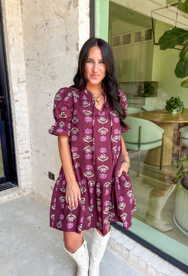 Rich fall tones meet sweet, feminine details in this burgundy mini dress. Covered in a soft pink floral print, it features a smocked finish for a flattering fit and short puff sleeves for just the right amount of charm. The ruffle-edged self-tie v-neckline adds a romantic touch, while side pockets keep it as practical as it is pretty. Made with a cotton blend for comfort and ease, this dress is your go-to for any special occasion!