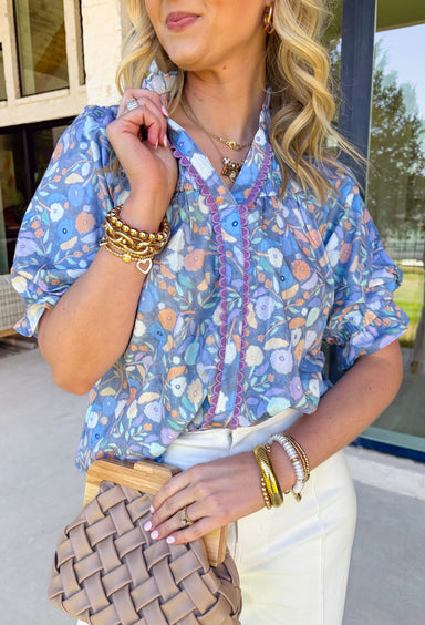 Soft, dreamy, and made for sunny spring days — this faded blue top features a delicate washed floral print in peach, airy blues, and light purple tones that feel effortlessly fresh. The vintage-inspired finish gives it that perfectly worn-in charm while still feeling polished and feminine. Light purple scalloped trim runs along the front for the sweetest pop of contrast, while subtle smocking adds texture and flattering shape. 