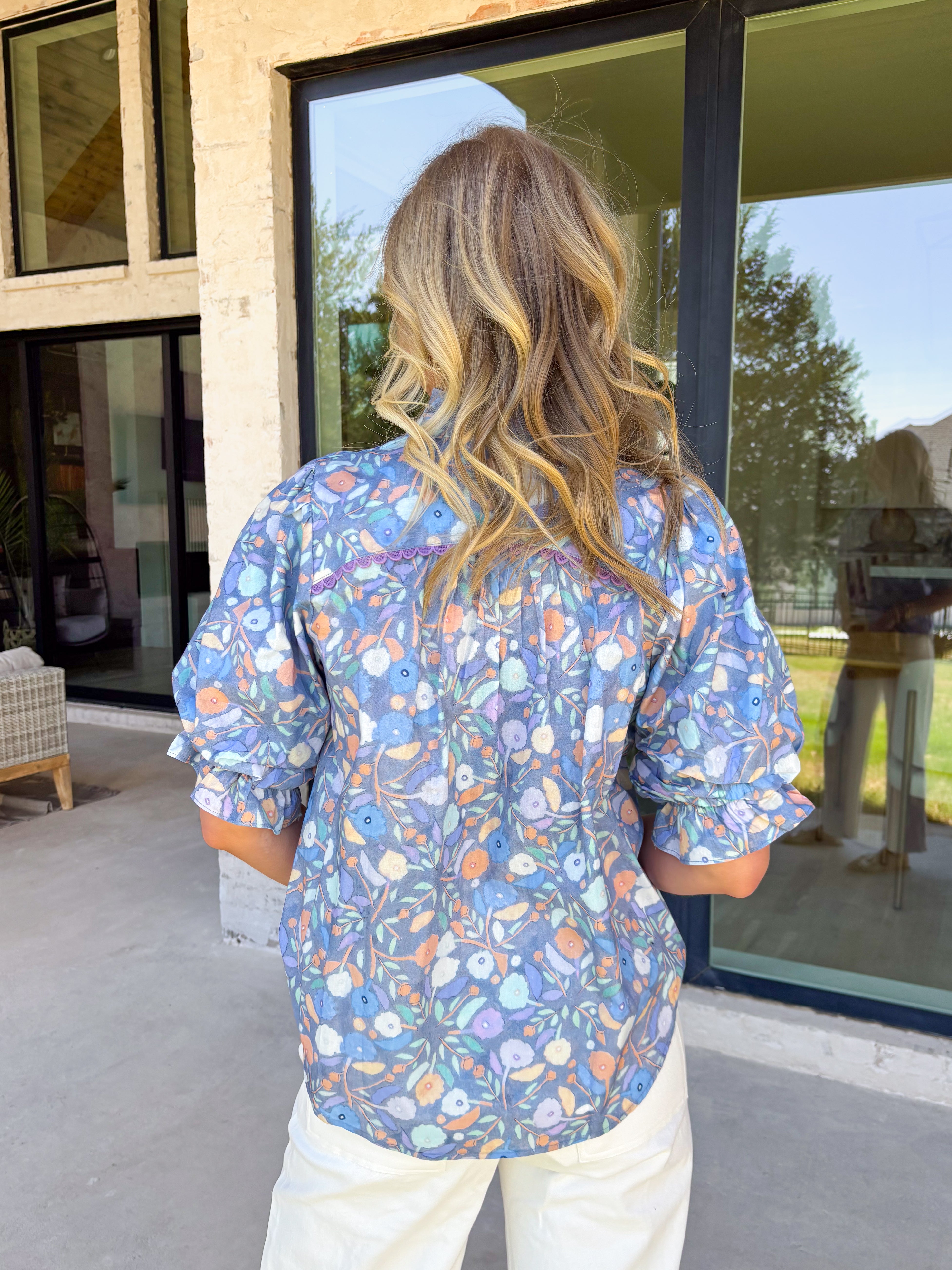 Soft, dreamy, and made for sunny spring days — this faded blue top features a delicate washed floral print in peach, airy blues, and light purple tones that feel effortlessly fresh. The vintage-inspired finish gives it that perfectly worn-in charm while still feeling polished and feminine. Light purple scalloped trim runs along the front for the sweetest pop of contrast, while subtle smocking adds texture and flattering shape. 
