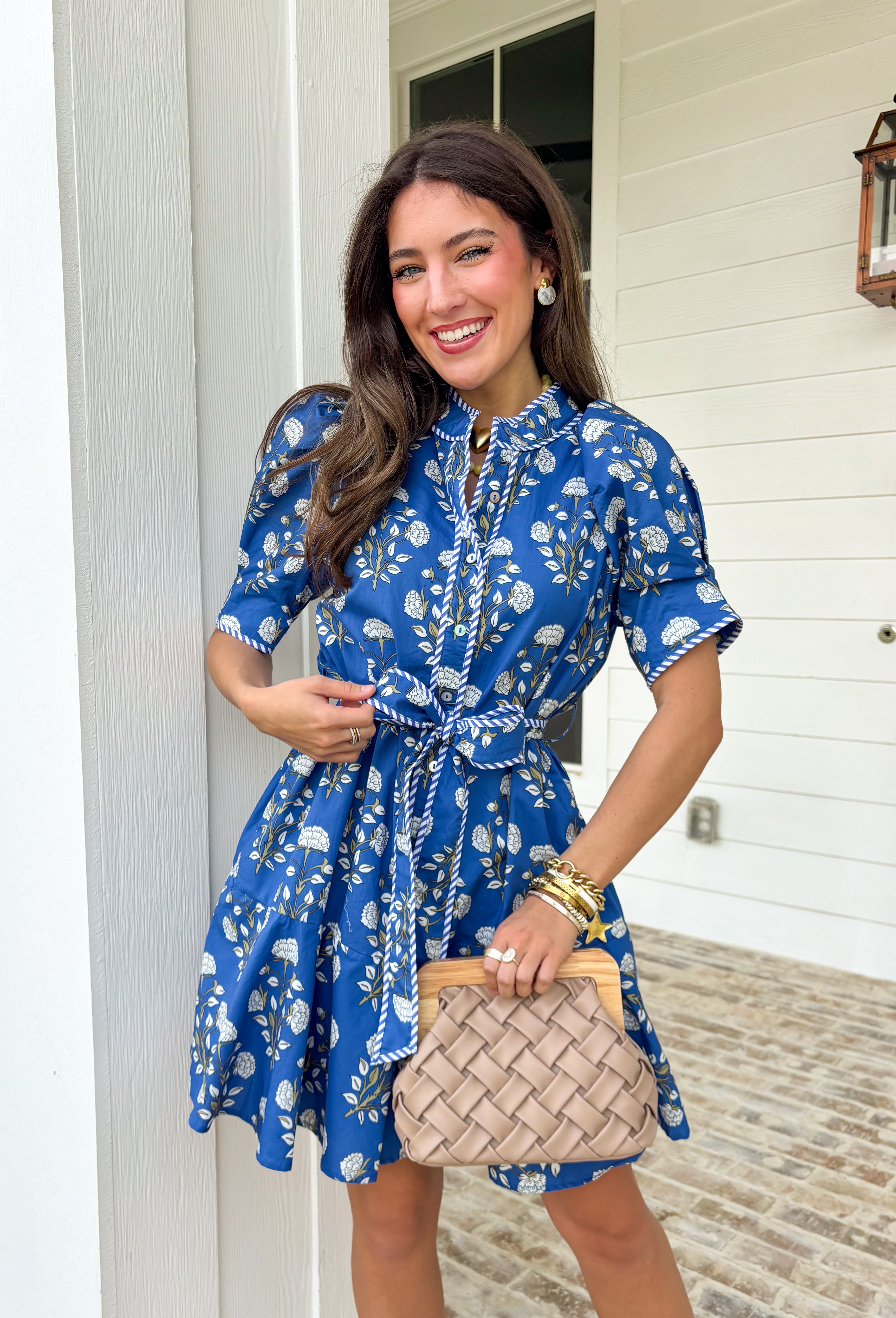 Fresh, fun, and full of spring charm, this blue short sleeve button-down dress by Entro is an easy standout. Featuring a floral print throughout, it brings the perfect pop of color while still feeling effortless and wearable. Blue and white striped trim accents the design for a playful contrast, and the matching self-tie waist gives it a flattering, adjustable fit. Finished with side pockets and a fully lined cotton feel, this dress is perfect for brunch, spring events, and sunny weekends.