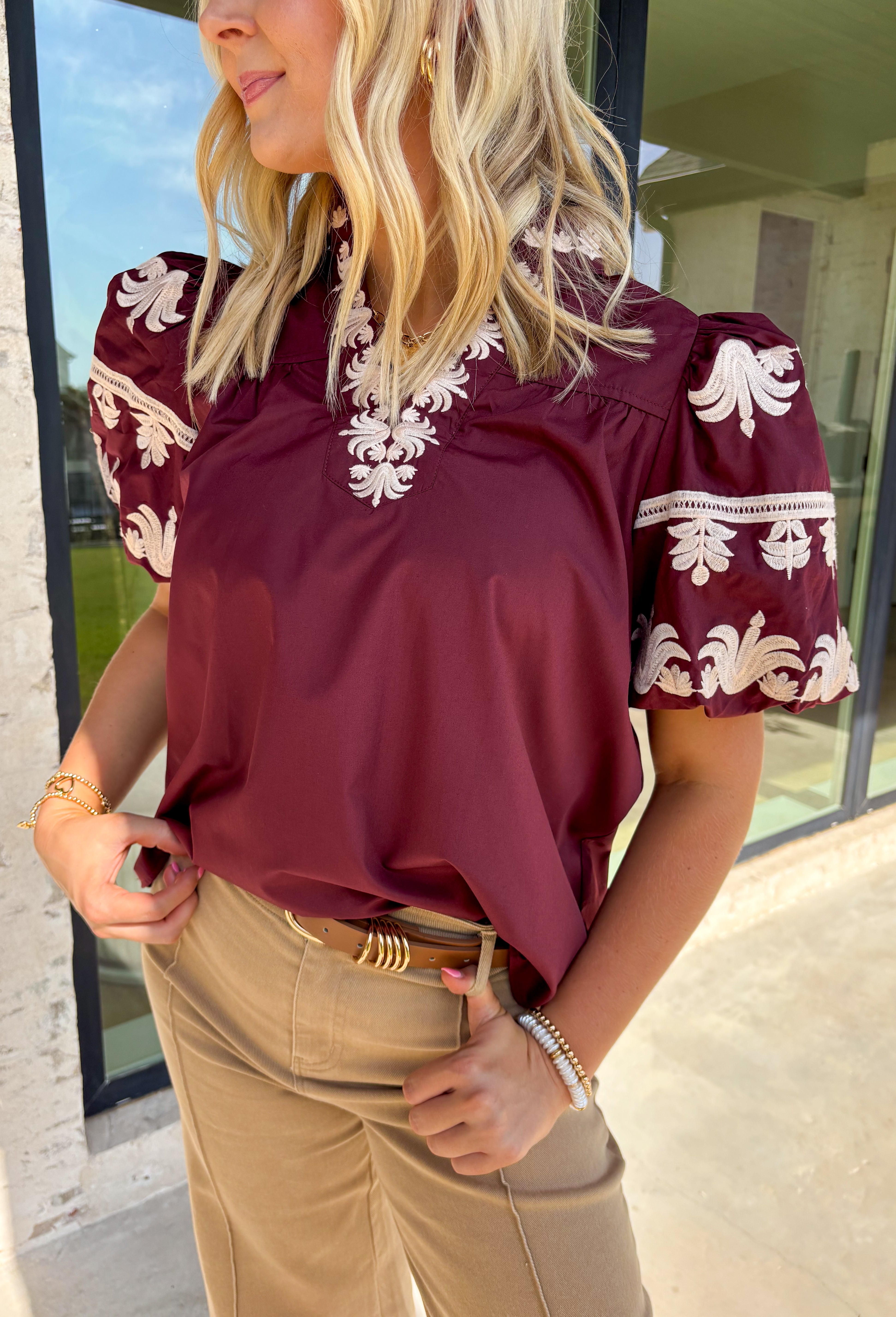 A rich autumn hue and elegant details make this burgundy top a seasonal standout. The v-neck band collar frames the neckline beautifully, while the embroidered placket adds a refined, textural touch. Short bubble sleeves bring a playful element to the silhouette, and the soft cotton-blend fabric keeps it comfortable from work to weekend. It’s the perfect piece to pair with denim, trousers, or skirts for a versatile fall look.