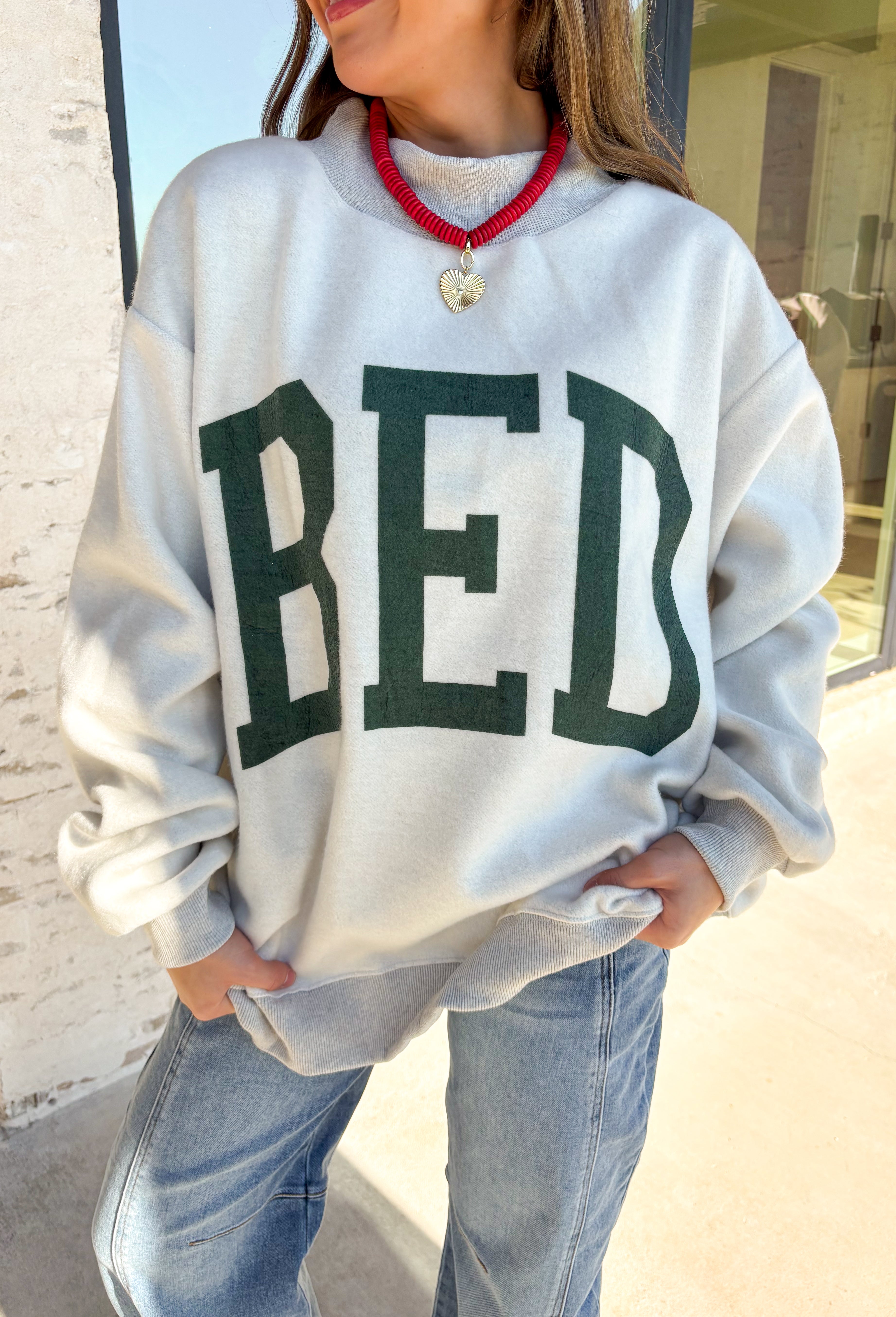 Two moods, one cozy pullover. This reversible mock neck sweater gives you options with light gray on one side and soft white fleece on the other. Printed in hunter green, one side reads “Gym” while the reverse says “Bed,” letting you wear your vibe for the day. With its oversized fit and cozy texture, it’s the perfect piece for lounging, layering, or heading out in casual comfort.